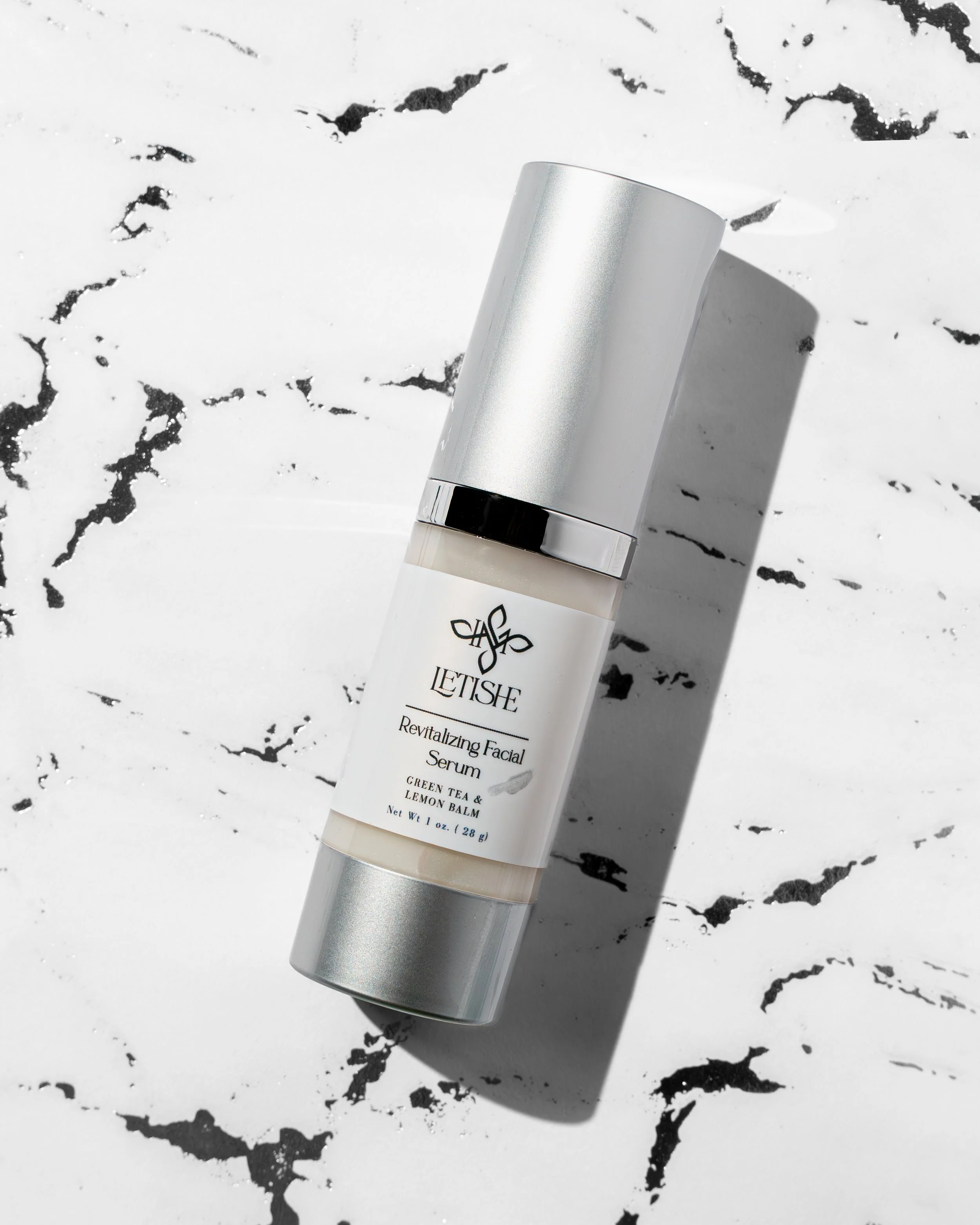 A white cylindrical bottle of LETISHE revitalizing facial serum with a silver cap on a white marble surface with black veining, casting a shadow.