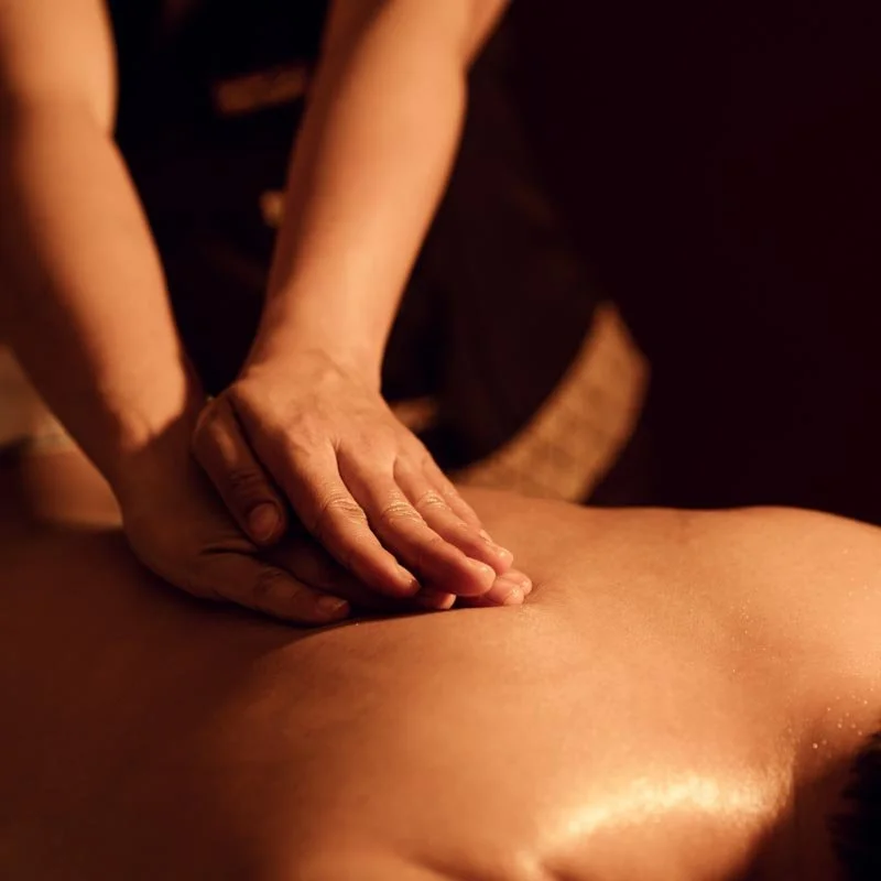 Close-up of a person receiving a massage, lying face down with a massage therapist's hands on their back, in a warm-lit environment.