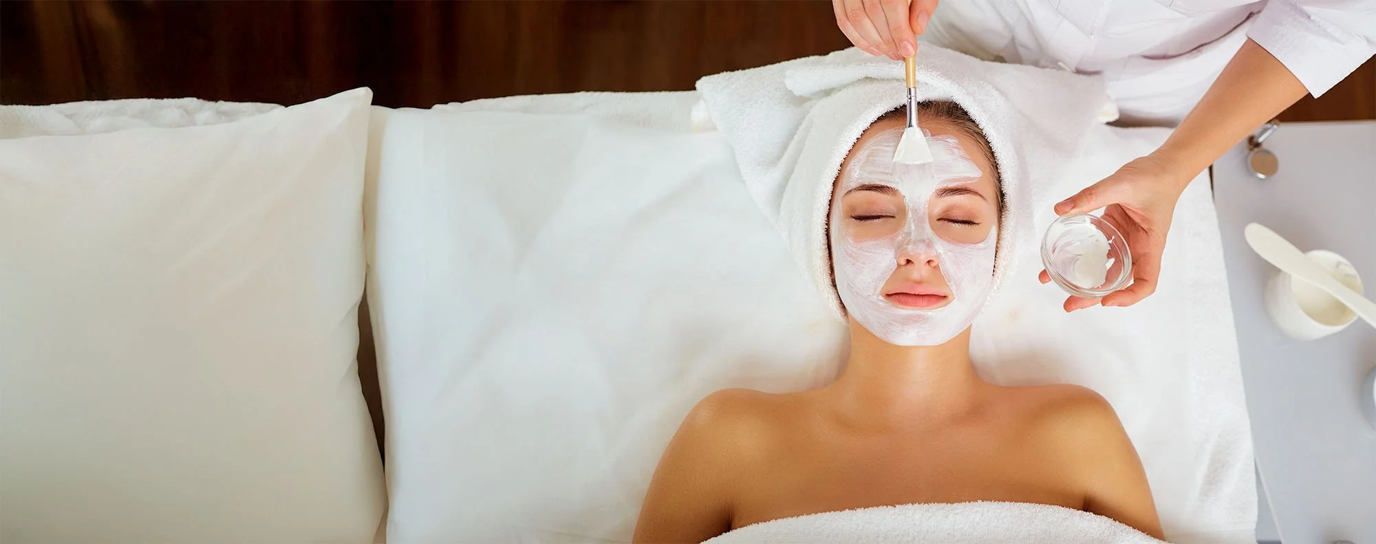 Woman receiving a facial treatment with a white face mask, lying on a bed with a towel wrapped around her head, in a spa or skincare clinic.