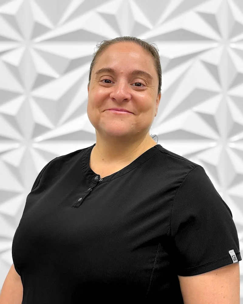 A woman with brown hair pulled back, wearing a black top, smiling, standing in front of a white geometric patterned wall.