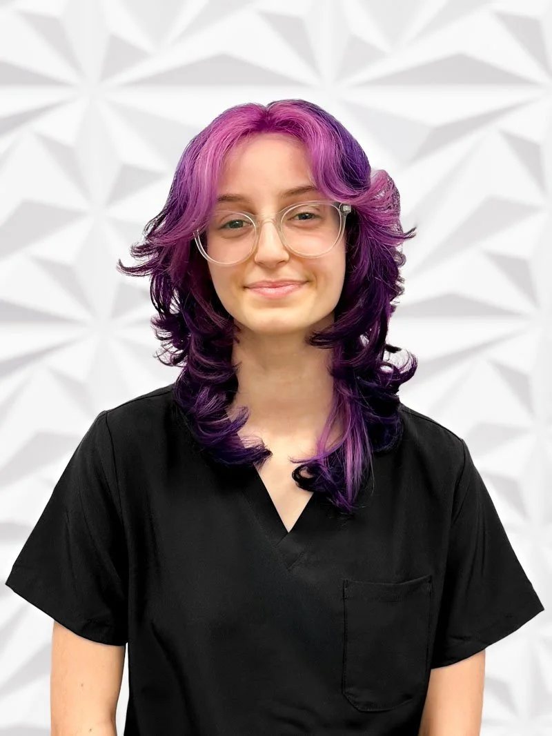 Young woman with purple and black curly hair wearing glasses and a black shirt, standing in front of a textured white wall.