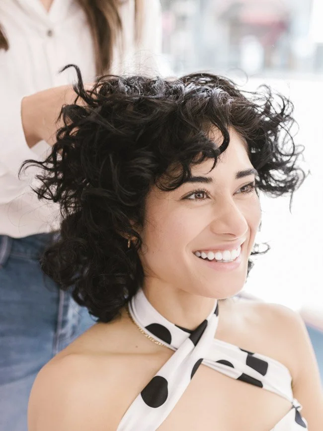 Woman with short curly dark hair smiling while getting her hair styled at a salon.