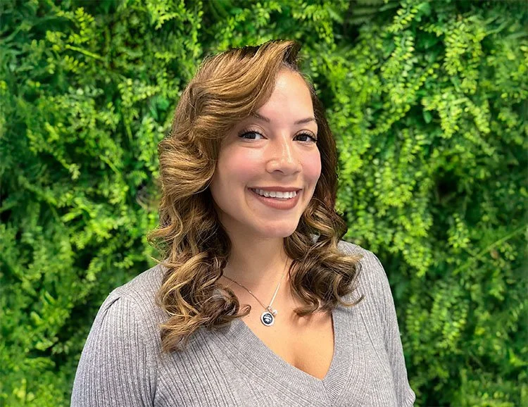 A woman with curly light brown hair and a bright smile, wearing a gray top and a necklace, standing outdoors in front of a lush green leafy background.