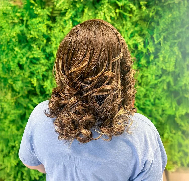 Back view of a person with shoulder-length, curly brown hair wearing a gray shirt against a green leafy background.
