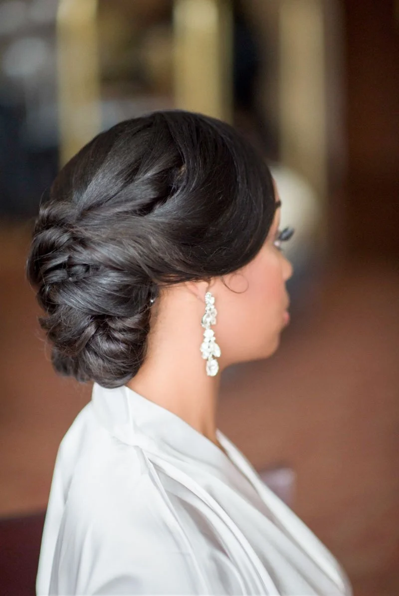 Side view of a woman with dark, styled hair in an elegant updo, wearing dangling earrings and a white satin robe.