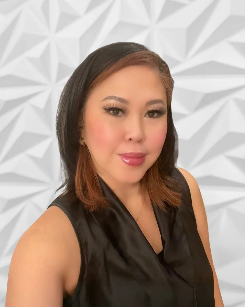 Portrait of a woman smiling, with long brown hair, wearing a black top, gold hoop earrings, and a gold necklace, standing in front of a textured white background.