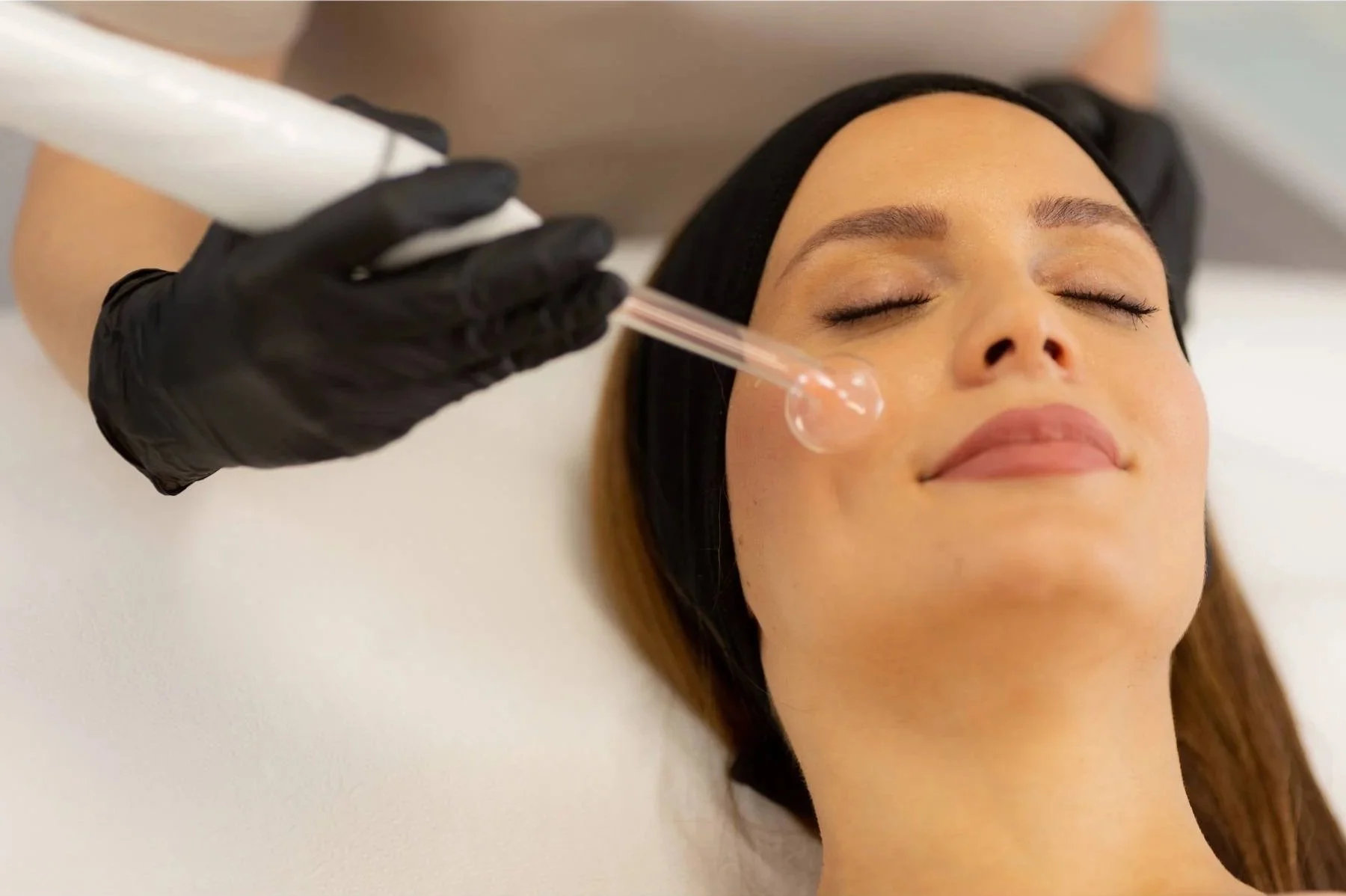 A woman receiving a facial treatment with a serum dropper, lying with eyes closed and a relaxed expression, in a spa or skincare clinic.