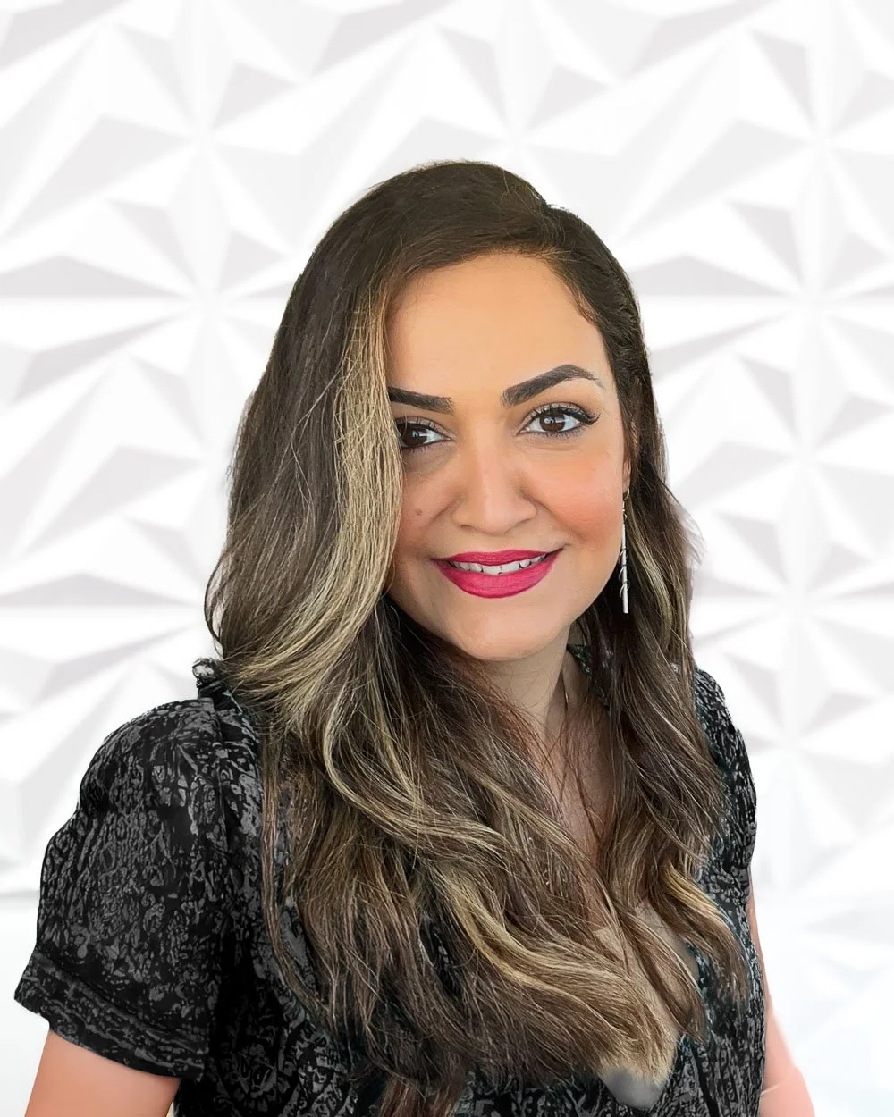 A woman with wavy brown hair and blonde highlights, smiling, wearing a black patterned top, against a white geometric background.