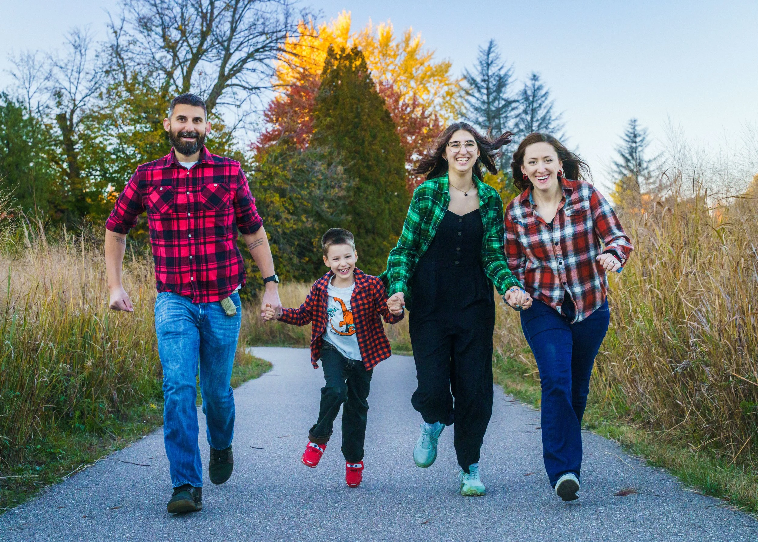A group of five people, three adults and two children, walking together on a paved path in a park during autumn. They are smiling and holding hands, with colorful fall trees in the background.