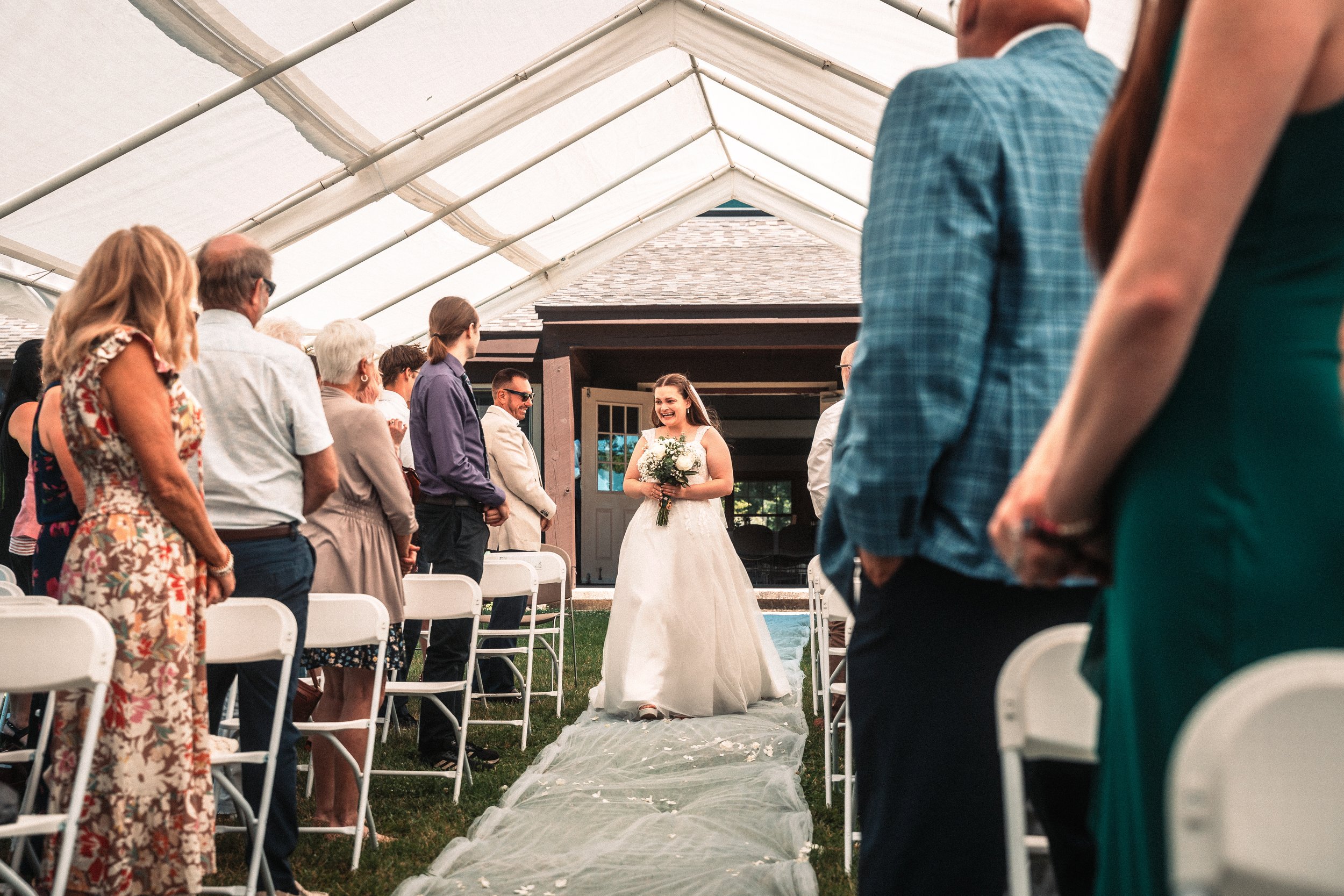A bride holding a bouquet is walking down the aisle at an outdoor wedding ceremony, with guests standing and watching in a tented setting.