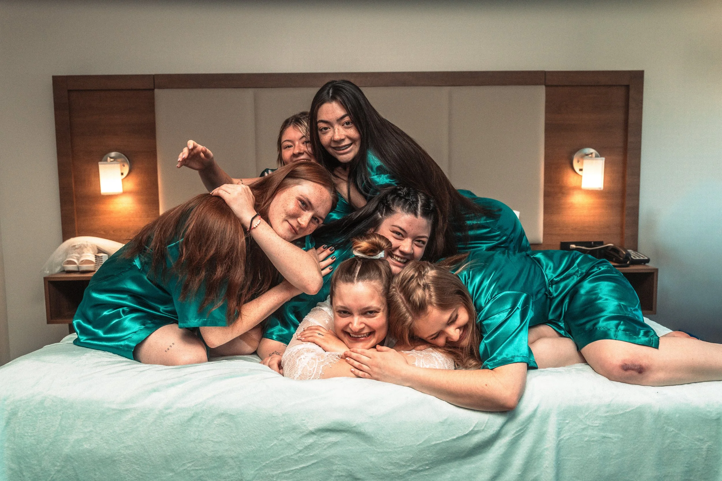 A group of young women in teal silky pajamas lying on a bed, smiling and hugging each other in a cozy hotel room.
