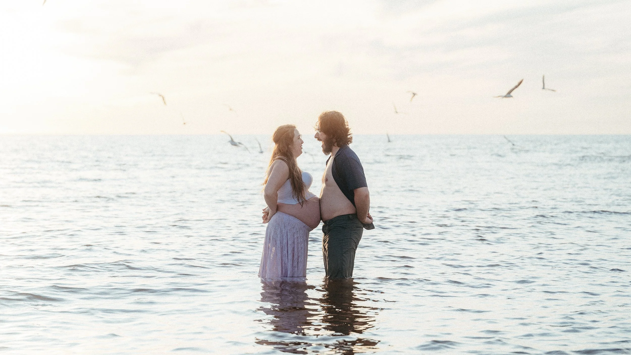 A pregnant woman and a man are standing in the ocean, facing each other and smiling, with seagulls flying in the sky behind them.