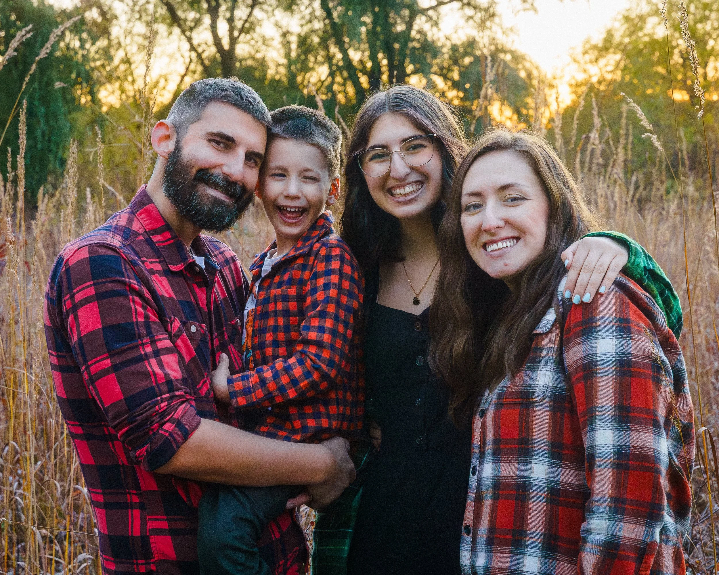 A family of four smiling and hugging in a field with tall grass, during sunset. The father and son are on the left, and the mother and daughter are on the right. All are wearing plaid shirts, with the family enjoying a warm, golden hour outdoors.