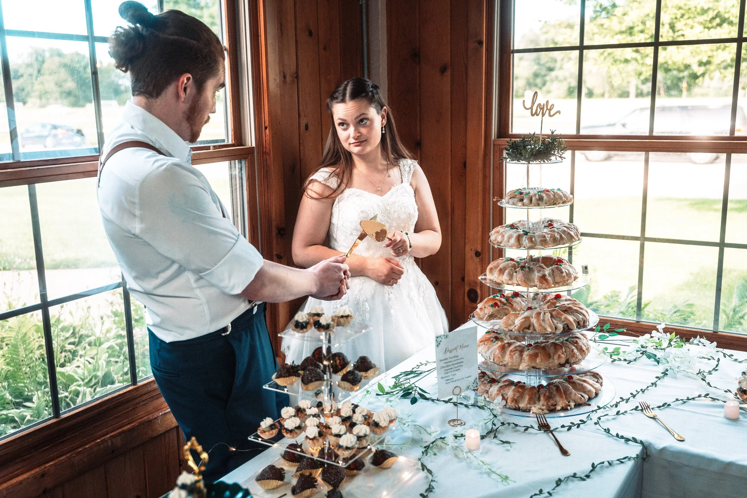 A bride and groom at a wedding reception, with the groom cutting a cake, and a table filled with assorted desserts, including Bundt cakes decorated with red and green candies, and cupcakes, inside a room with large windows and wooden walls.