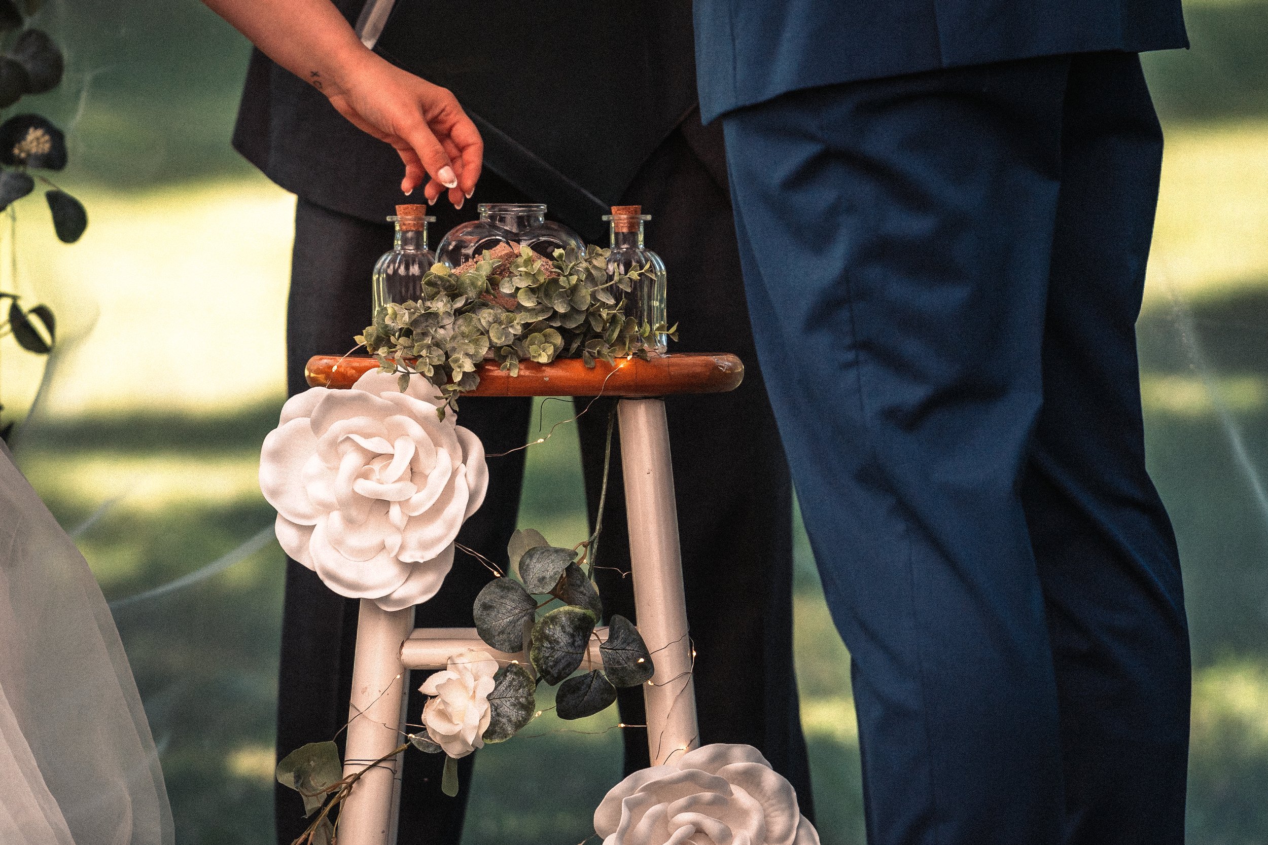 A person in a dark suit is holding a small bottle of glue and decorating a small wooden table with green vines, white flowers, and small glass bottles, in an outdoor setting with greenery in the background.