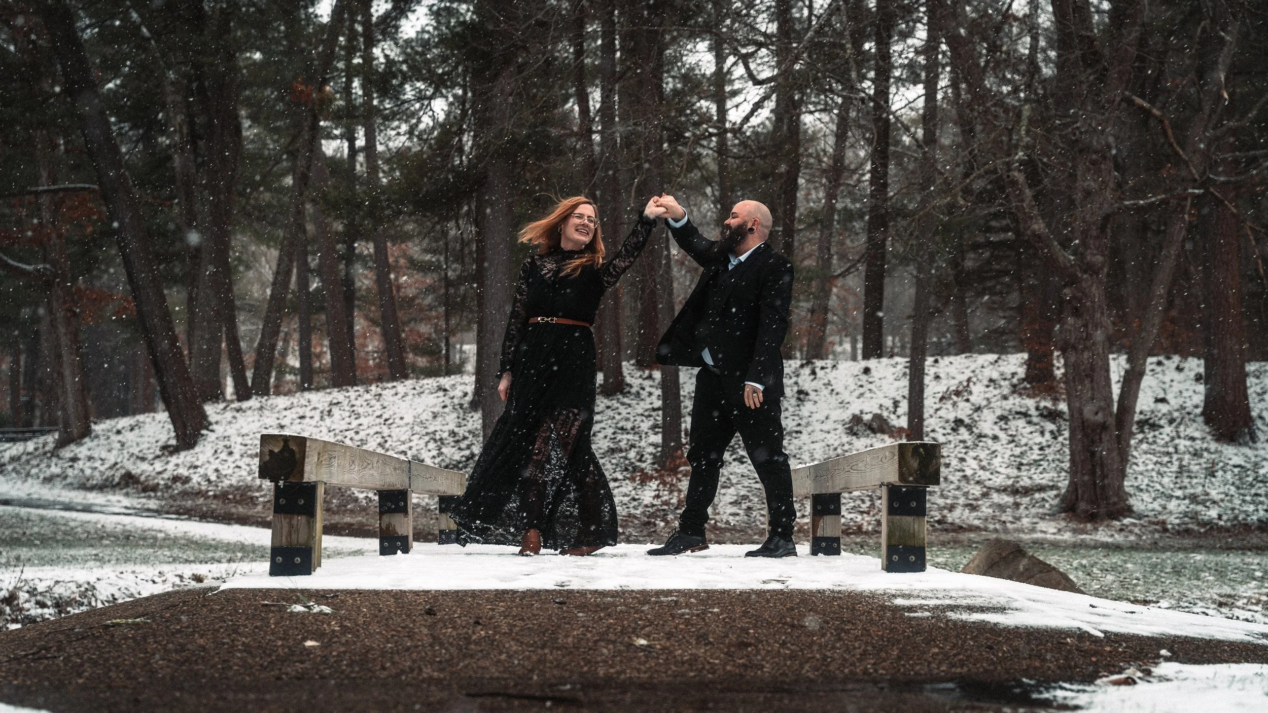 A couple in formal attire dancing and playing with snow on a small wooden bridge in a snowy wooded area. Photography by Kai Bass Photo.