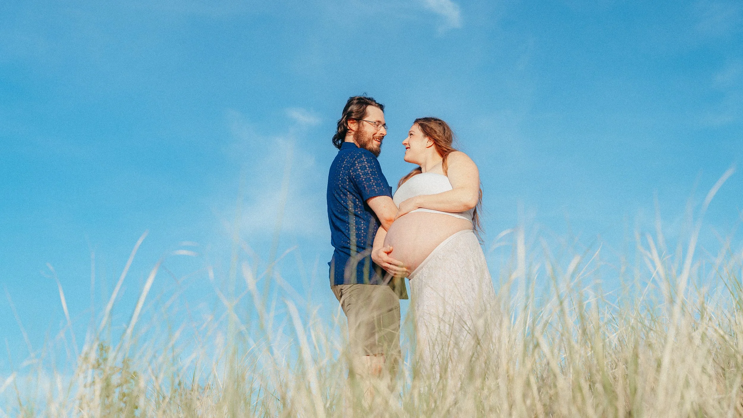 A happy expecting couple standing in a field under a blue sky, with the woman cradling her pregnant belly and the man embracing her.