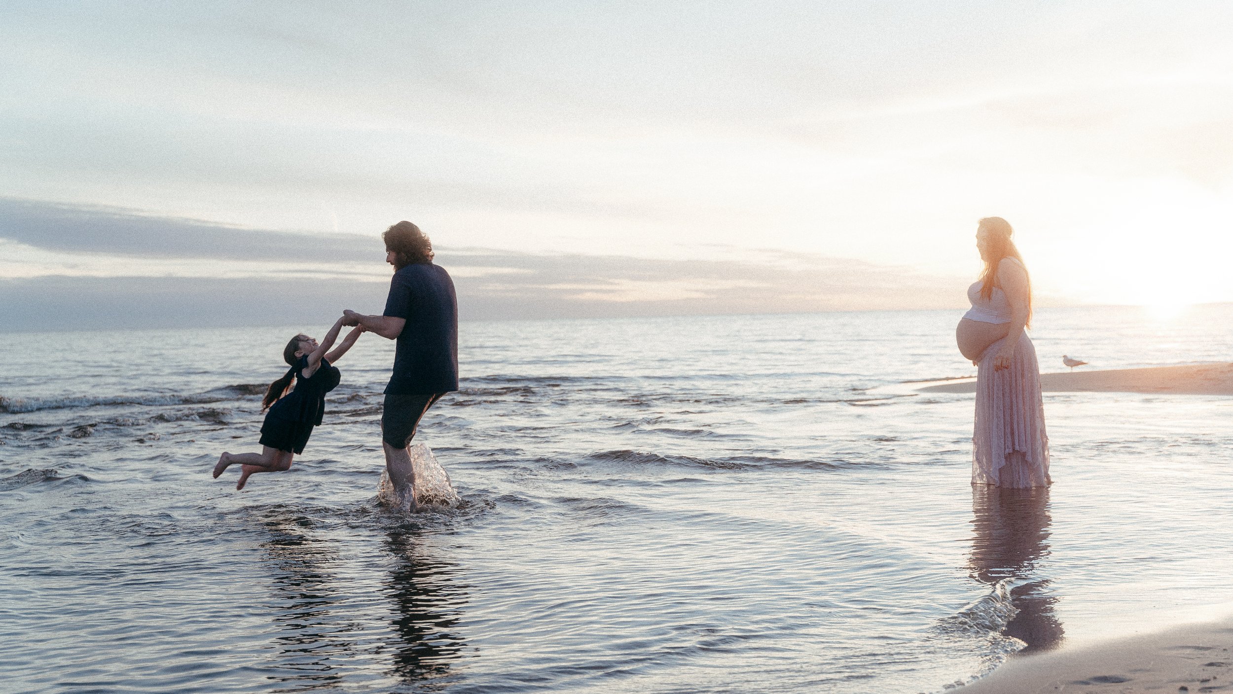 A family at the beach during sunset: a man is holding a girl and lifting her, a woman who appears to be pregnant stands nearby watching.