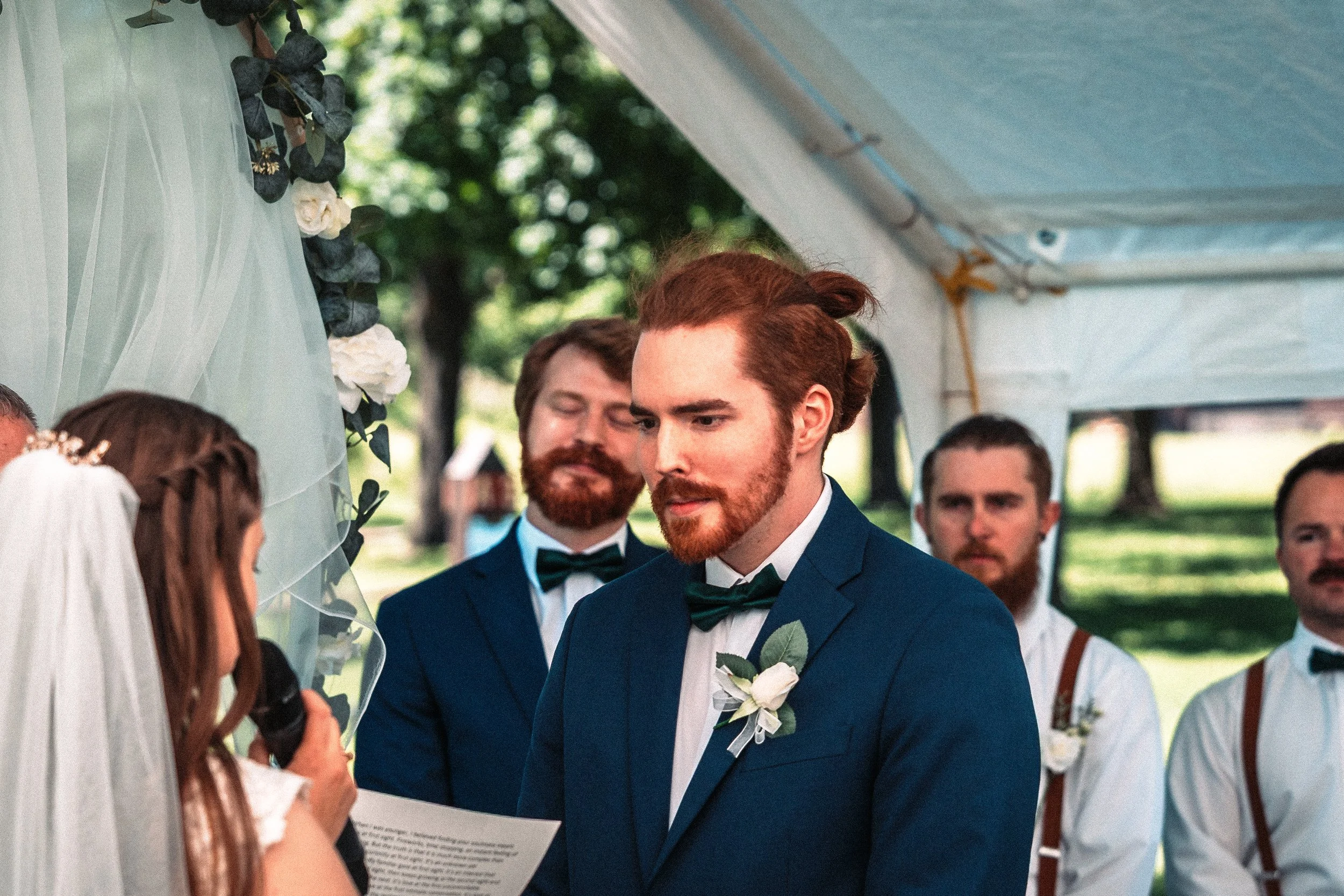 Groom and groomsmens standing in a wedding ceremony outdoors, exchanging vows under a decorated canopy with greenery and white flowers.