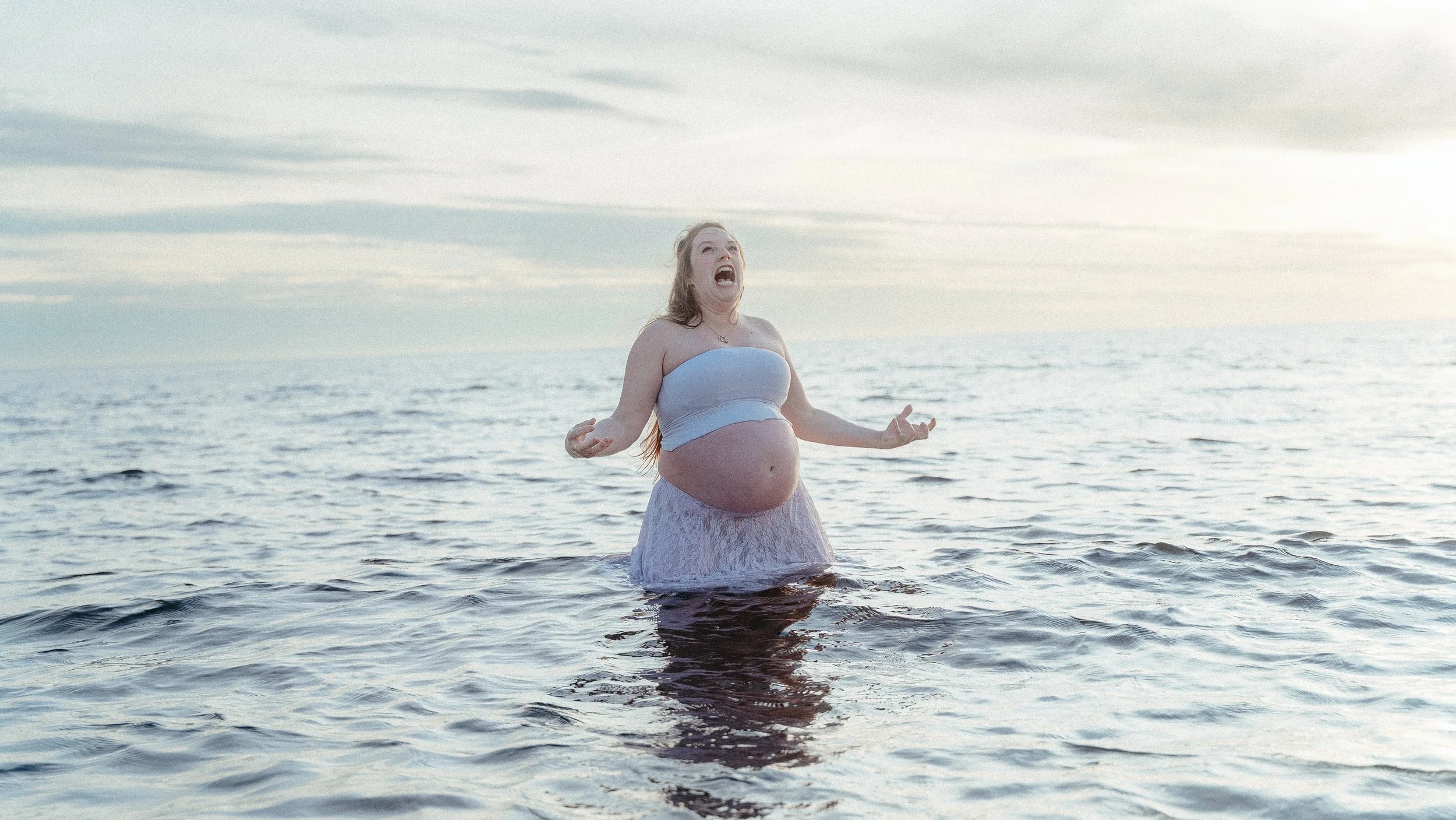 A pregnant woman in a strapless top and a lace skirt standing in the ocean during sunset, with her arms raised and her head tilted back making an expressive face.