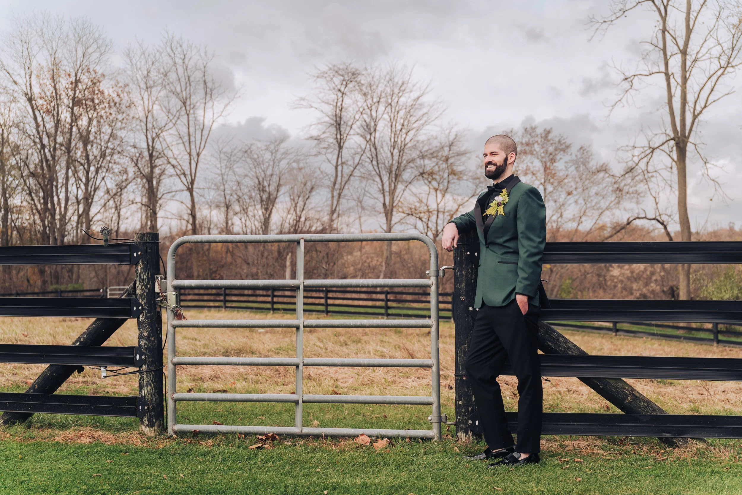 A man in a dark green tuxedo with black lapels and a floral boutonniere, standing outdoors by a black wooden fence and a metal gate, smiling amidst leafless trees and overcast sky at  riverbend estate wedding venue near Otsego, Michigan