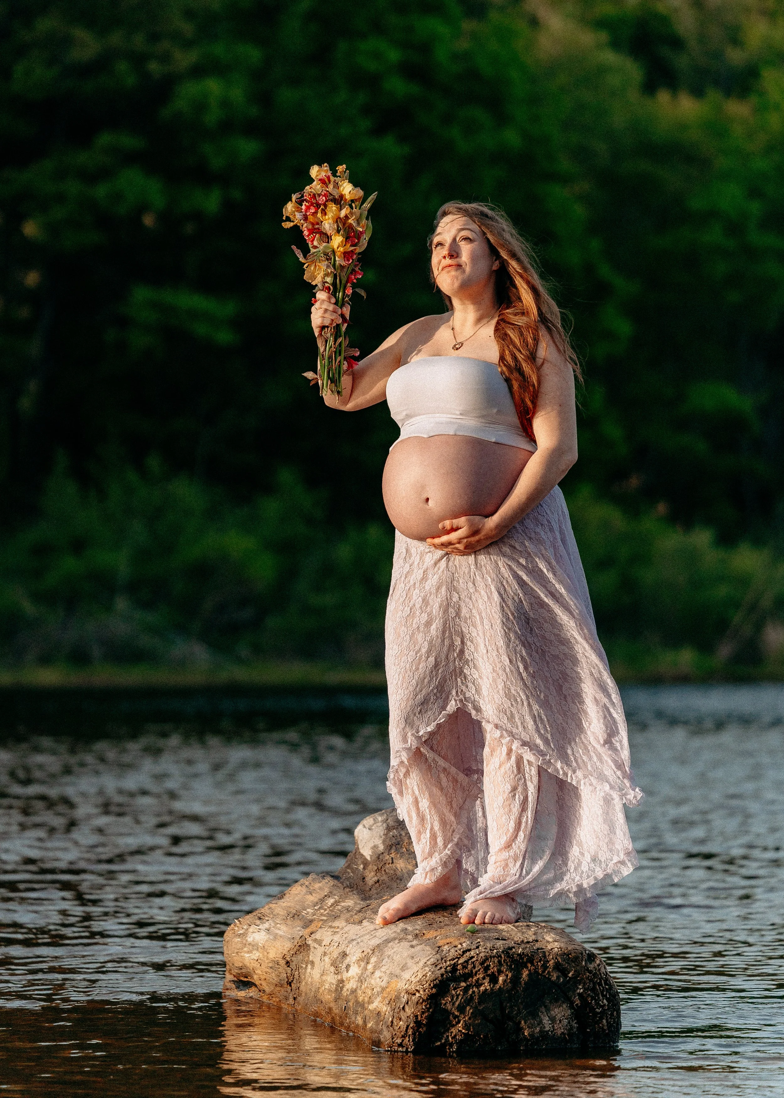 A pregnant woman with long red hair stands barefoot on a rock in a body of water, holding a bouquet of flowers. She is wearing a strapless top and a long, flowing skirt, and looks upward with a serene expression. The background features lush green tr