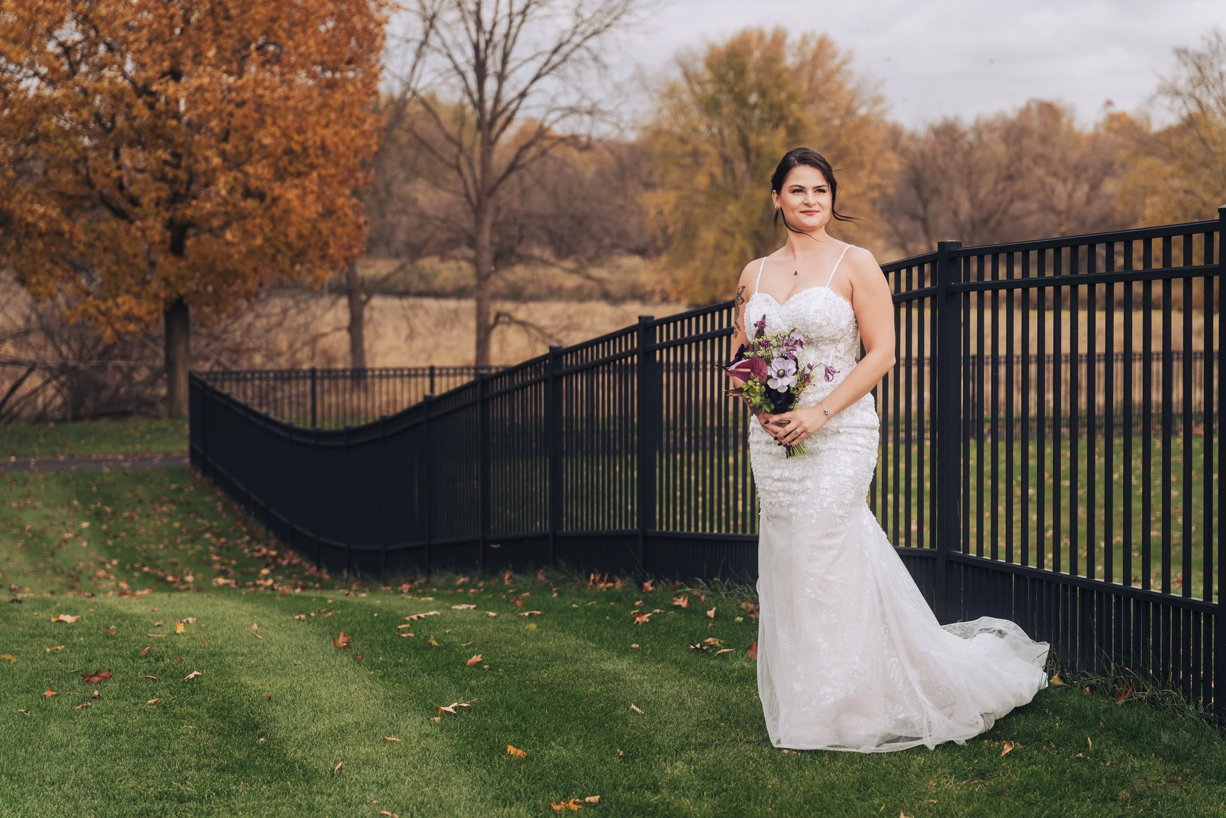 A bride in a white lace wedding dress holding a bouquet of purple, white, and pink flowers outdoors during autumn, standing near a black metal fence with trees and fall foliage in the background of riverbend estate wedding venue near Otsego, Michigan