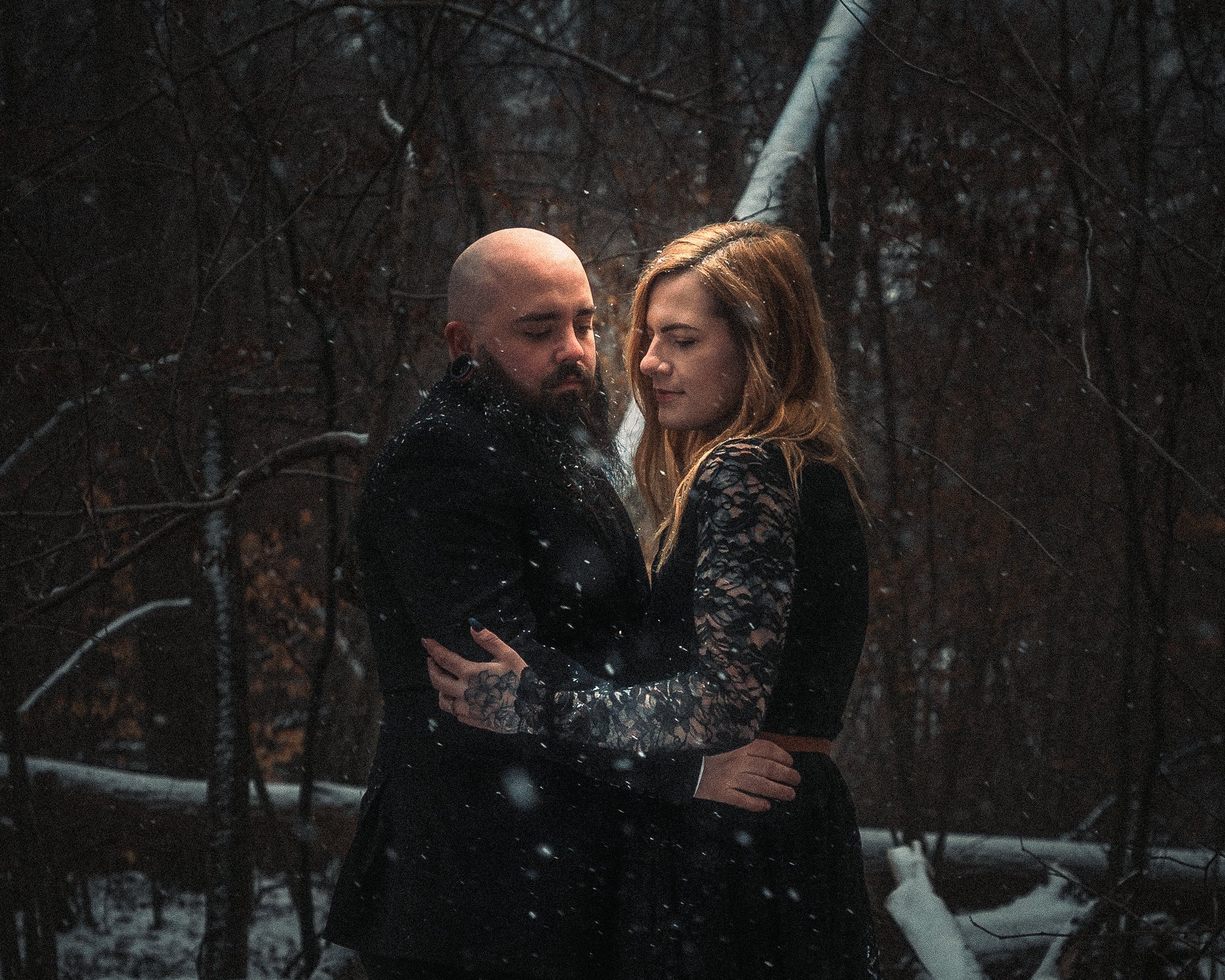 An alternative couple embracing each other in a dark, snowy forest with trees and a fallen tree in the background. Photography by Kai Bass Photo.