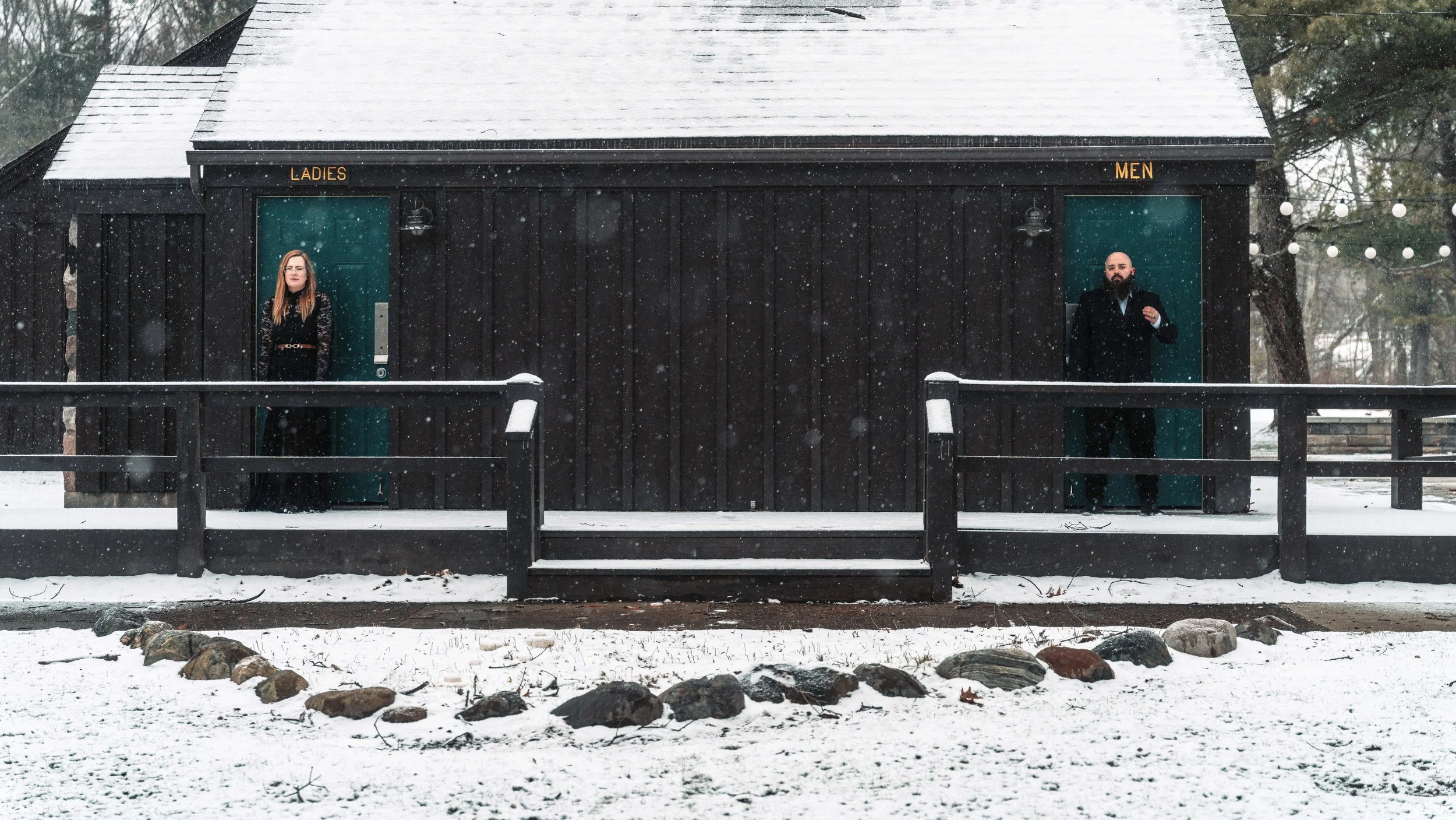A small black wooden building with two separate doorways labeled 'Ladies' and 'Men' on a snowy day. Two people, a woman on the left and a man on the right, are standing outside each door. Snow is falling, and there is a rock border in front of the bu