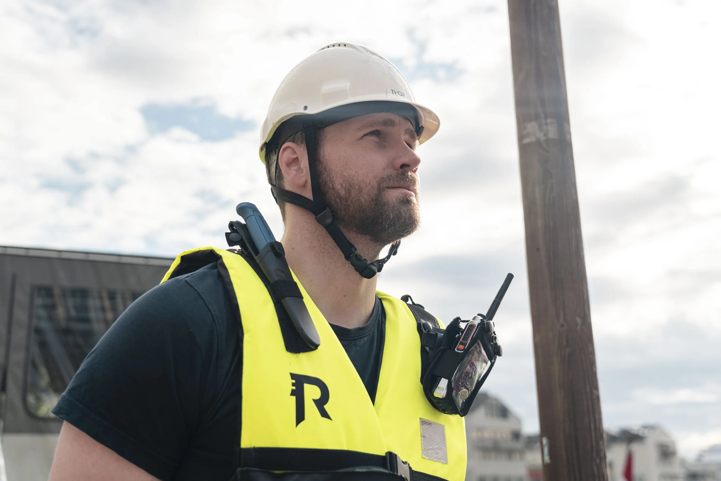 En mann iført hjelm, refleksvest og radiosender står ute, med elektrisk stolpe og bygninger i bakgrunnen.