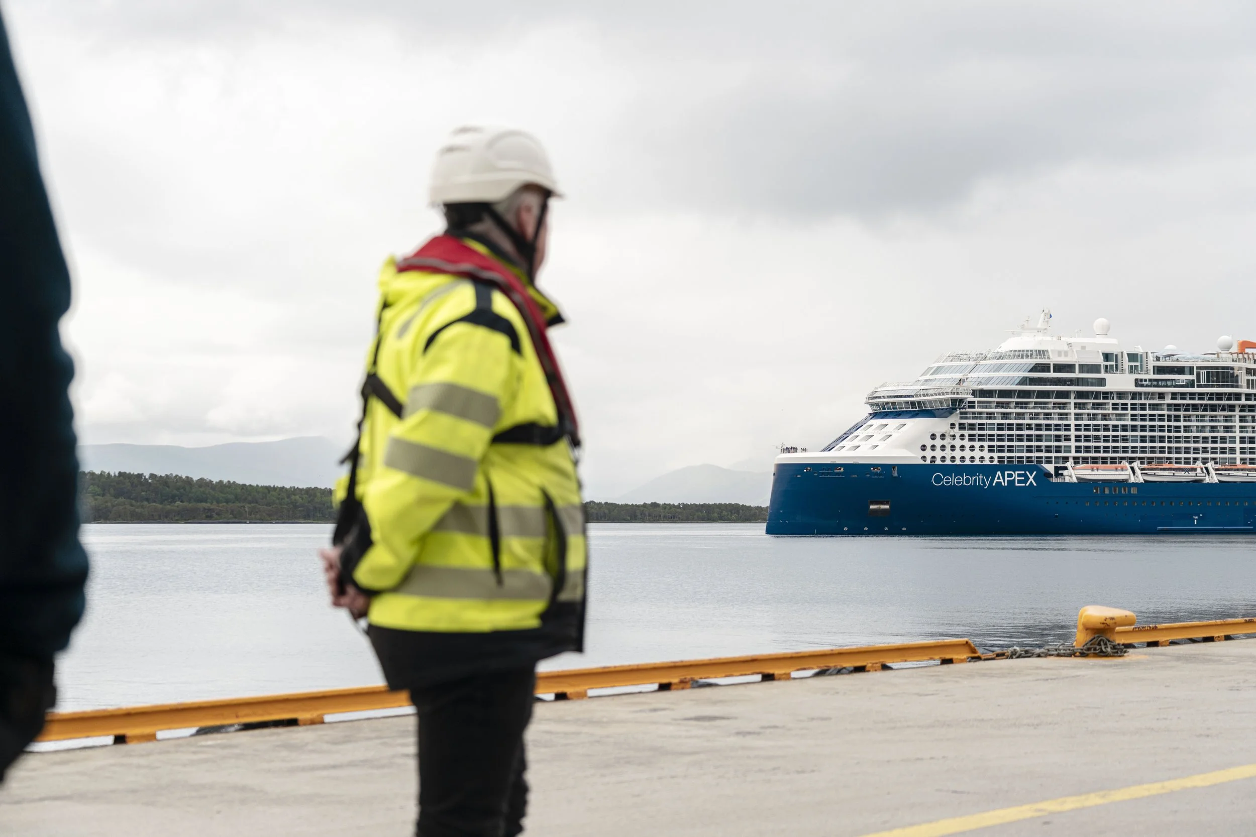 Personer i refleksvester og hjelm som står på kai med et stort cruiseskip i bakgrunnen, omgitt av vann og en overskyet himmel.