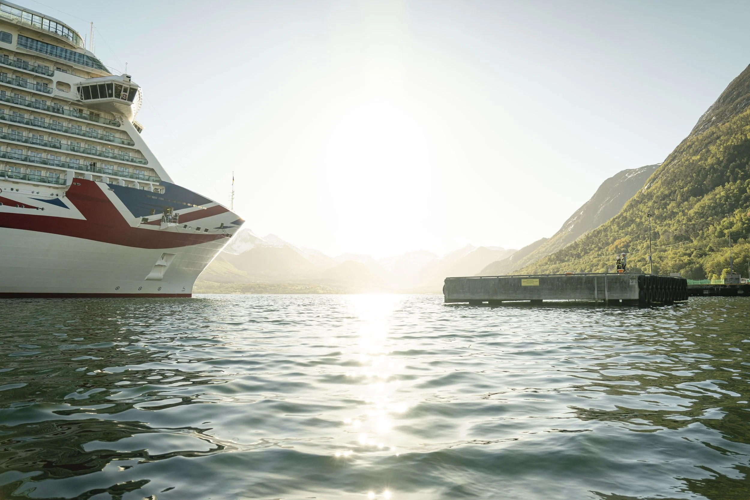 En stor cruiseferge ligger i en fjord omgitt av grønne fjell, med solen som skinner over vannet.