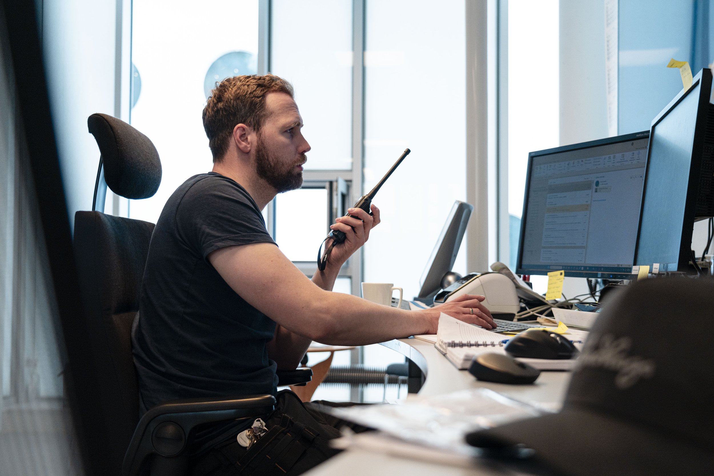 En mann med brunt hår og skjegg, iført svart t-skjorte, sitter ved et kontorbord og snakker i en radio. Han er omgitt av dataskjermer, papirer og kontorrekvisita.