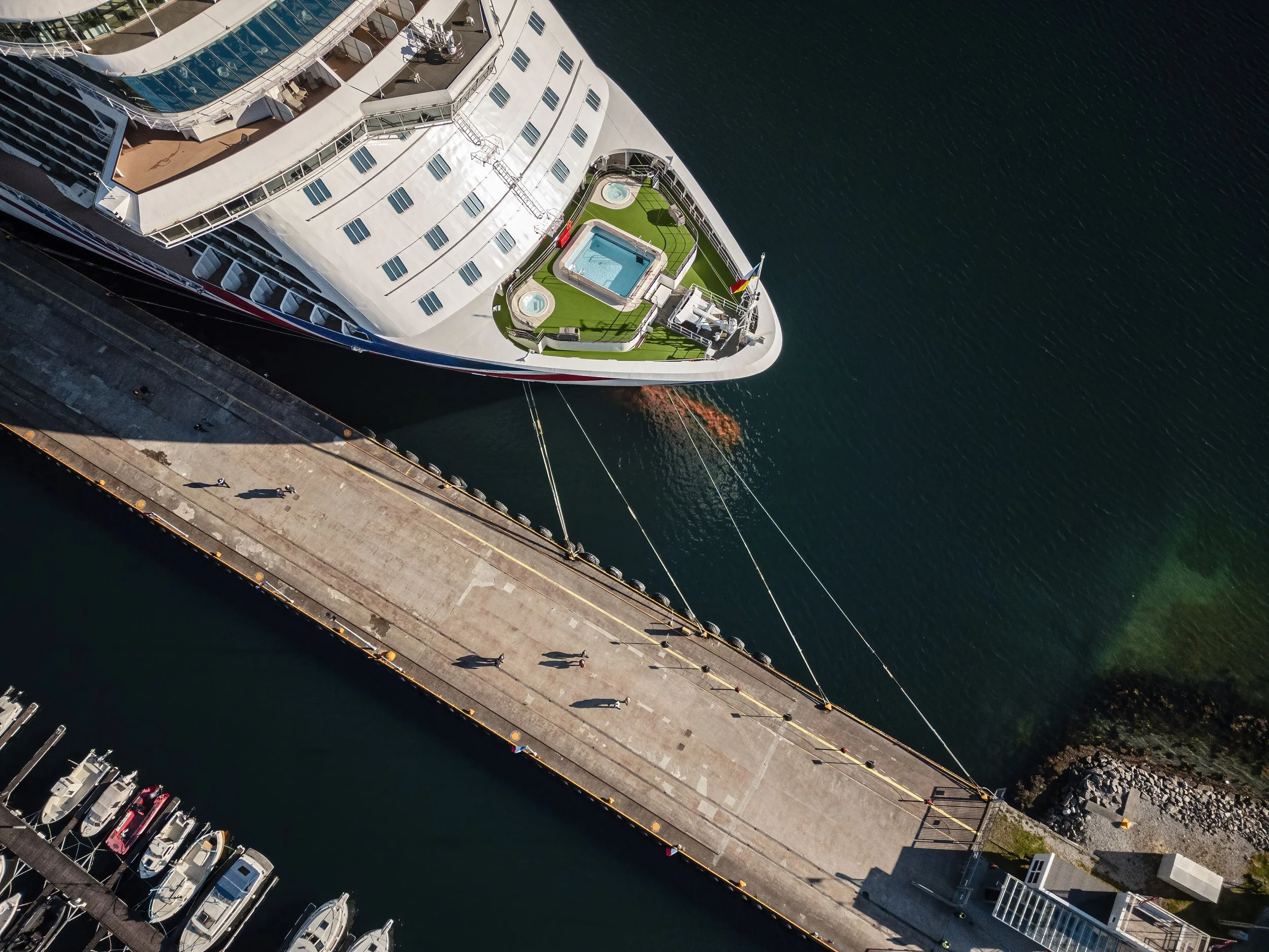 En stor hvit cruise-skip er dokket opp ved en kai. På toppen av skipet er det en solterrasse med to boblebad, kunstgress og noen sittegrupper.
