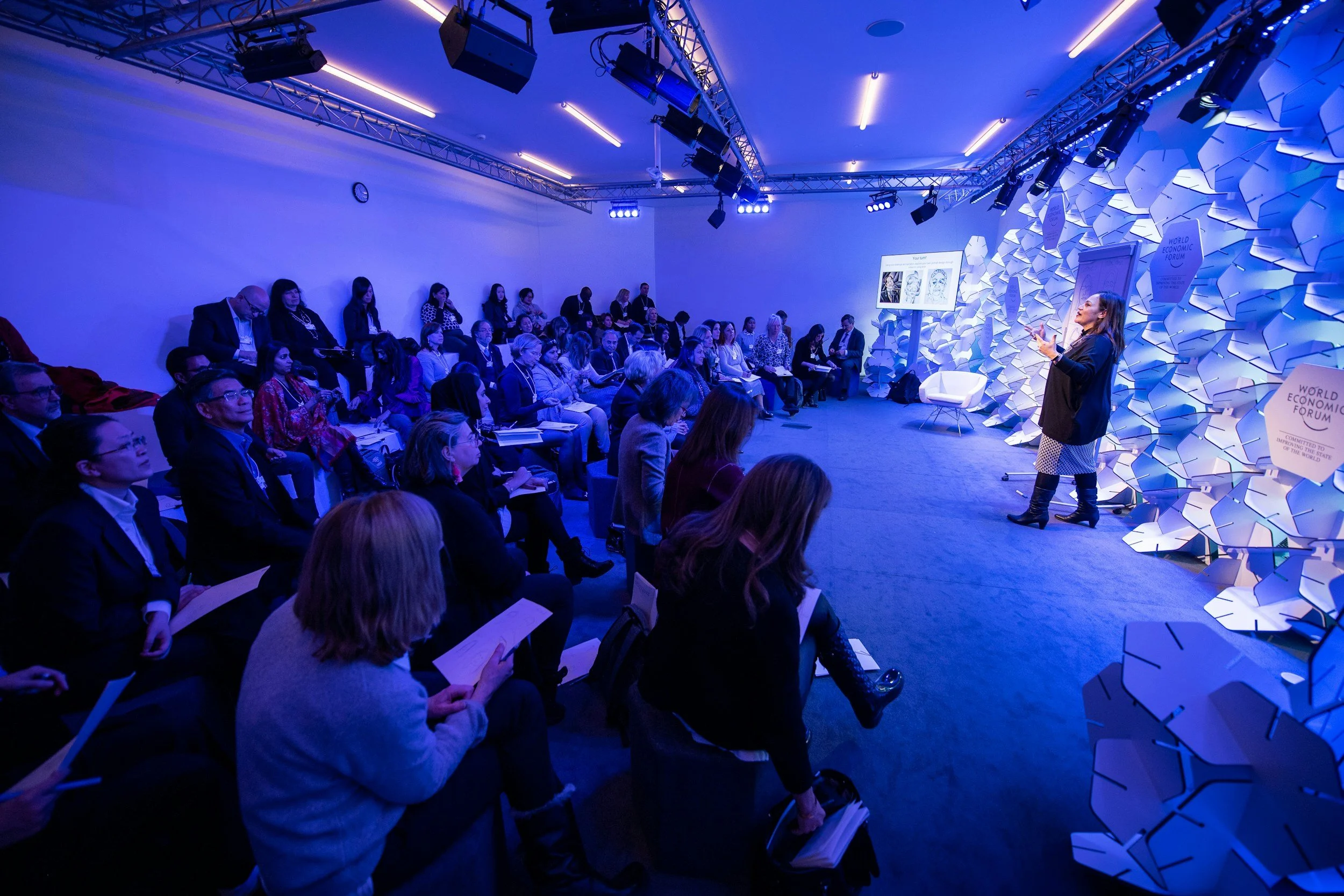 A speaker presenting to an audience at the World Economic Forum, in a room with blue lighting, modern wall decor, and overhead lighting fixtures.