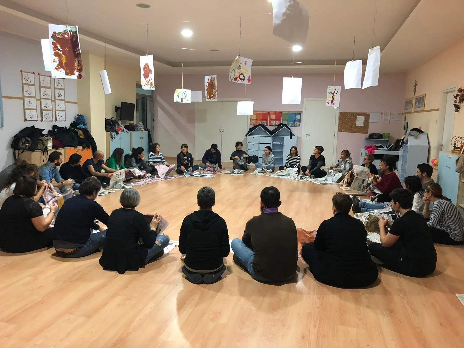 Gruppo di persone in un momento di corso di ludicità consapevole sedute in cerchio su un pavimento di legno, in un ambiente decorato con disegni e materiali artistici appesi al soffitto.