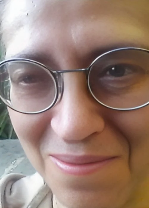 Una persona sorridente con occhiali tondi e capelli castani, che guarda verso la fotocamera.