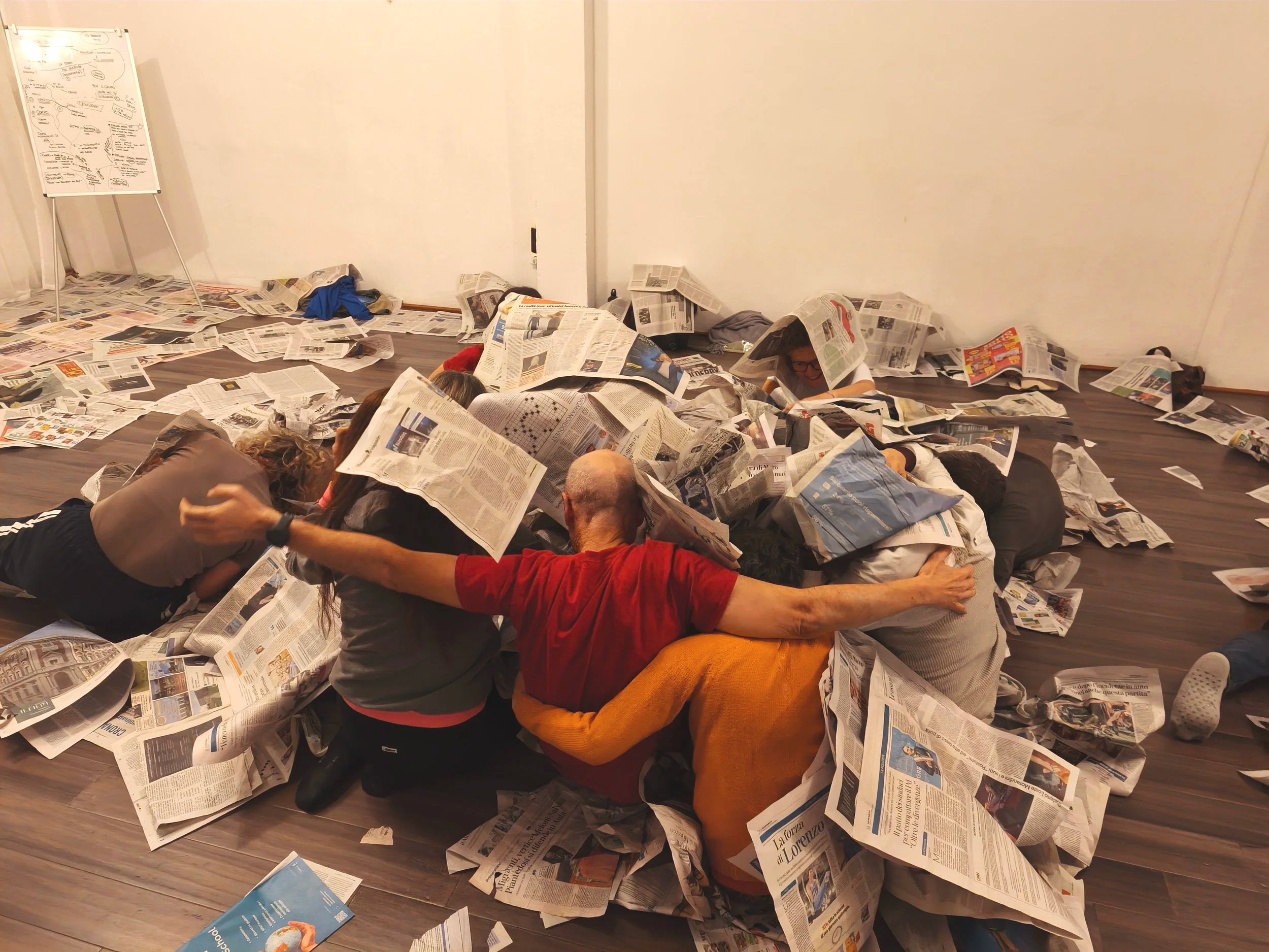 Gruppo di persone che si abbracciano e si concentrano sotto fogli di giornale sovrapposti, ambientato in una stanza in un momento di corso di ludicità consapevole