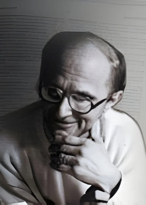 Ritratto in bianco e nero di un uomo con capelli radi e occhiali, che sorride o fa una smorfia, con la mano sotto il mento.