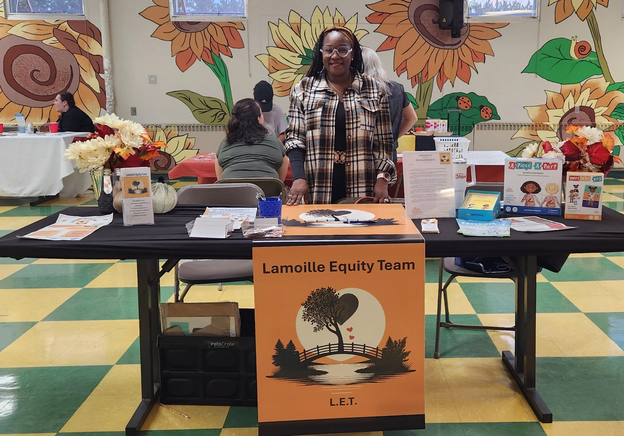 A woman standing behind a promotional table with a sign reading 'Lamoille Equity Team L.E.T.' in a room with colorful sunflower mural walls. The table has informational materials and decorations including flowers and pumpkins.