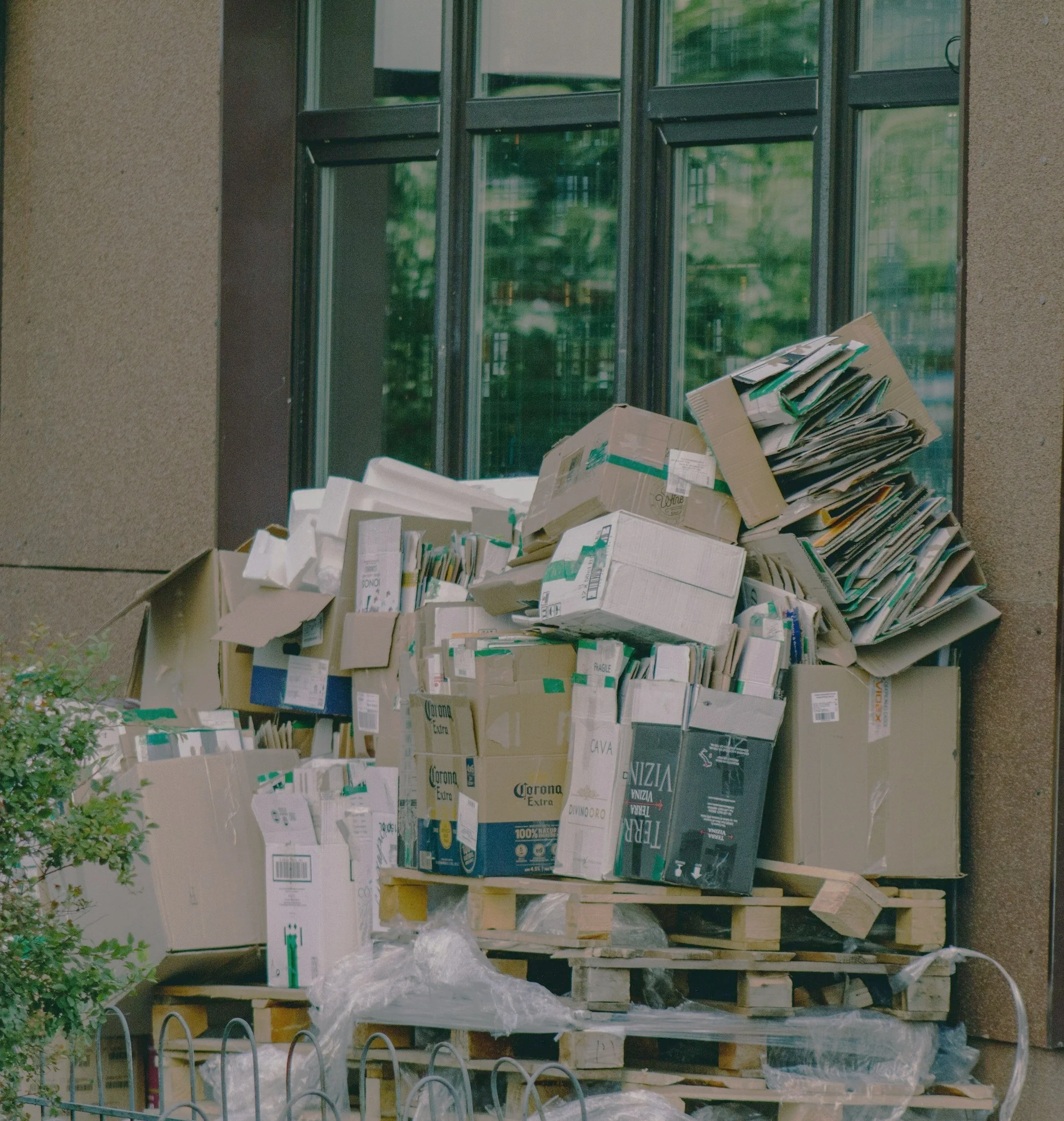 A large pile of cardboard boxes and flattened cartons stacked on wooden pallets outside near a building with large windows.