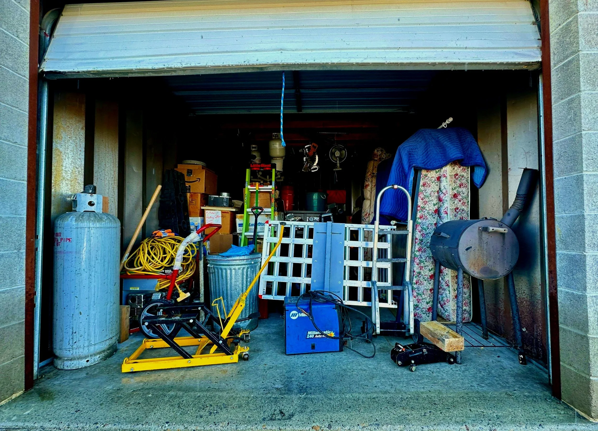 Inside a cluttered garage with various boxes, tools, and equipment, including a propane tank, a ladder, and a yellow hand truck.