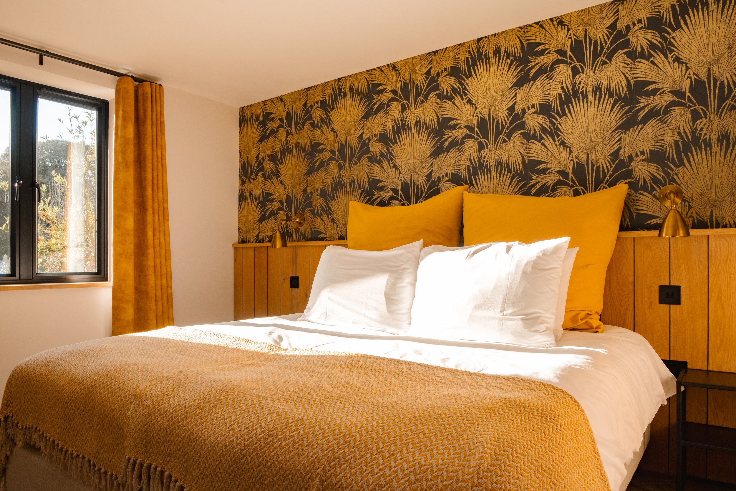 A neatly made bed with white sheets and pillows, two large mustard-yellow pillows, set against a dark wall with a gold leaf pattern. There are two small brass wall-mounted lamps on either side of the bed. A window with black framing and mustard-yello