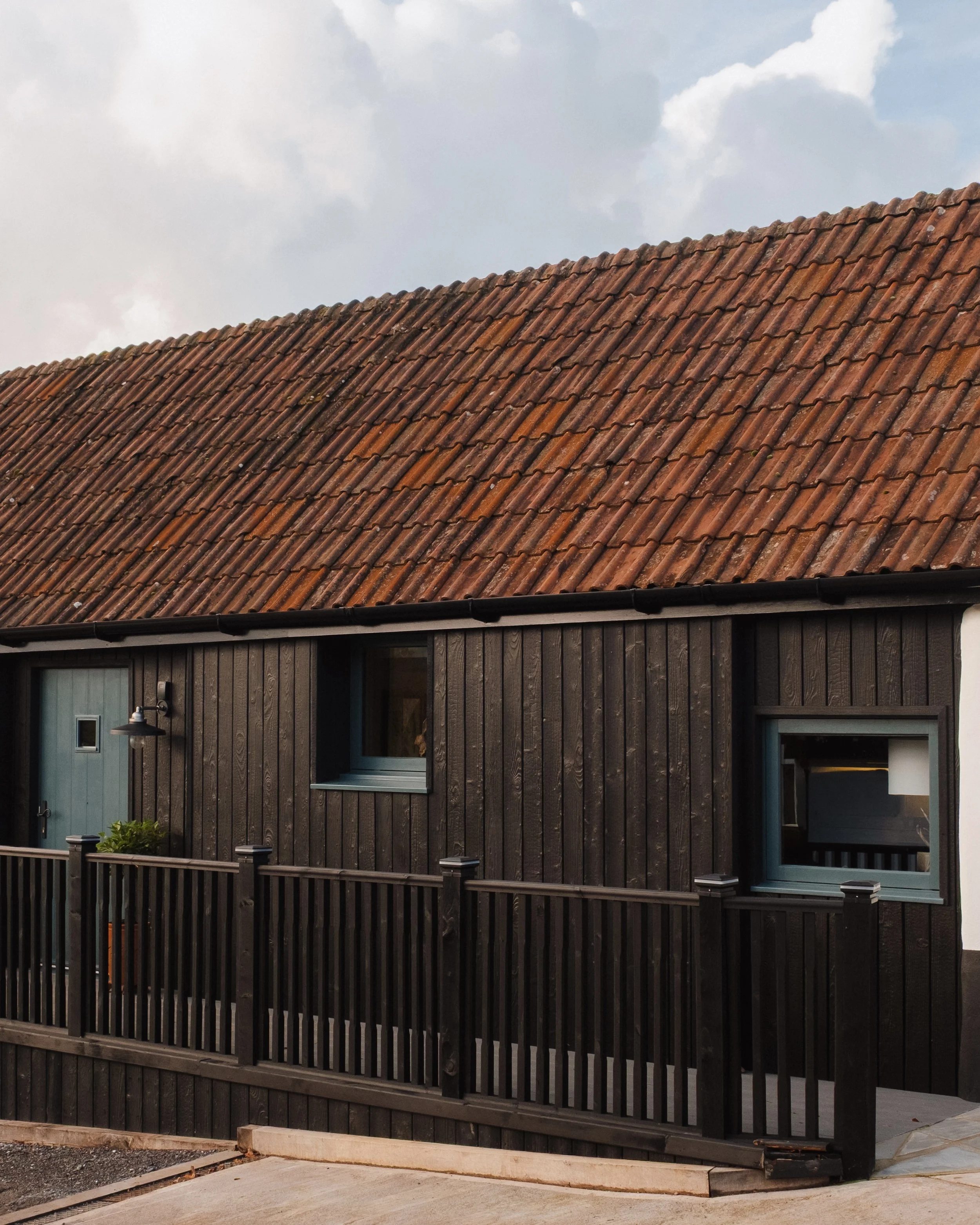 A house with black wooden siding, a black fence, and a red-tiled roof under a cloudy sky.