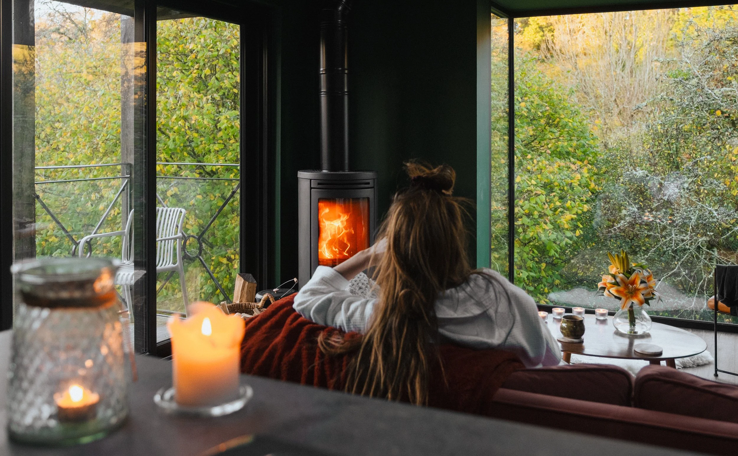 A woman with long hair, sitting on a red couch, looking at a wood-burning stove in a cozy living room with large windows overlooking green trees.