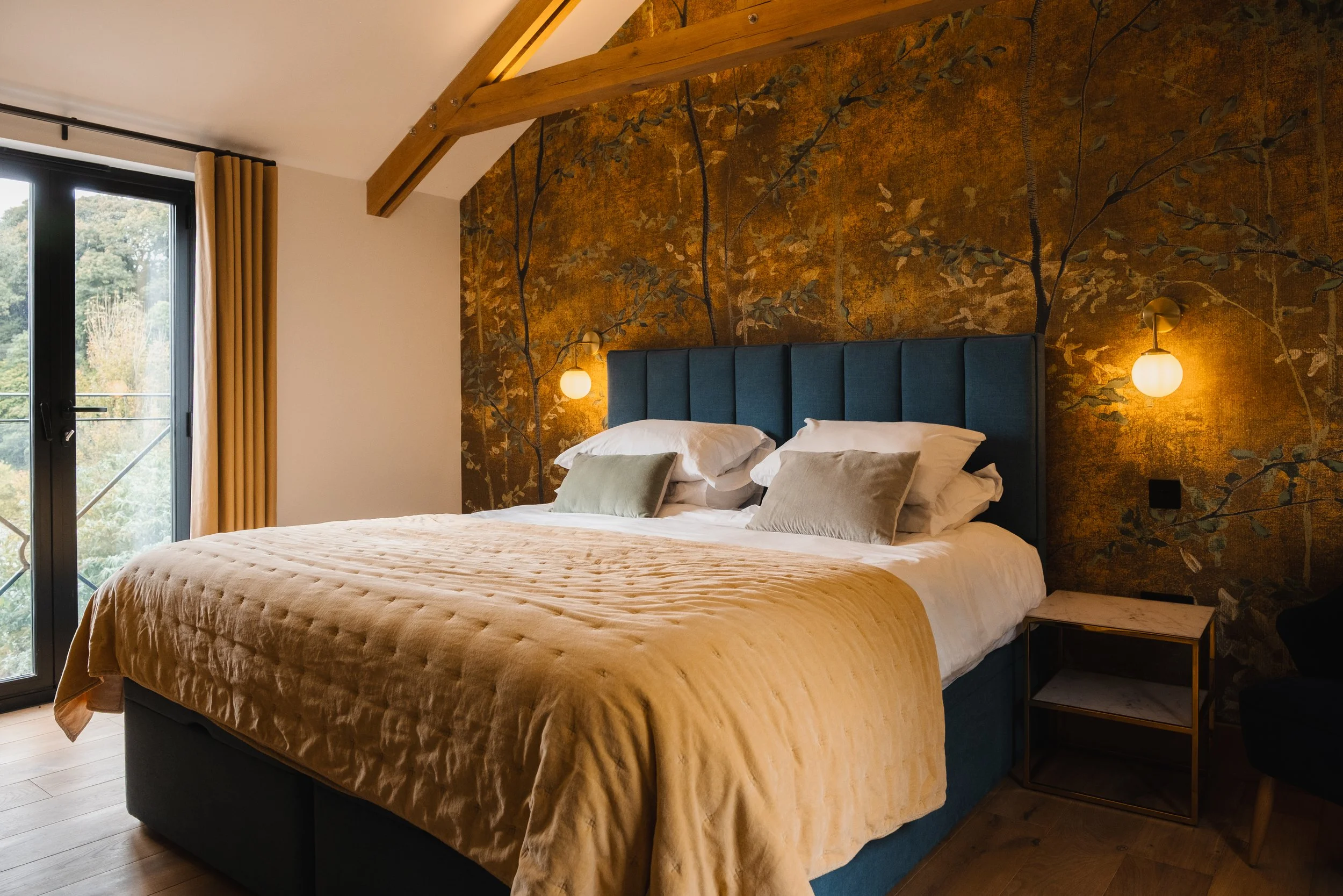 A neatly made bed with a blue upholstered headboard in a bedroom with wooden beams and a gold and brown floral wallpaper. There are two wall-mounted lights on either side of the bed, a small side table next to it, and large windows with beige curtain