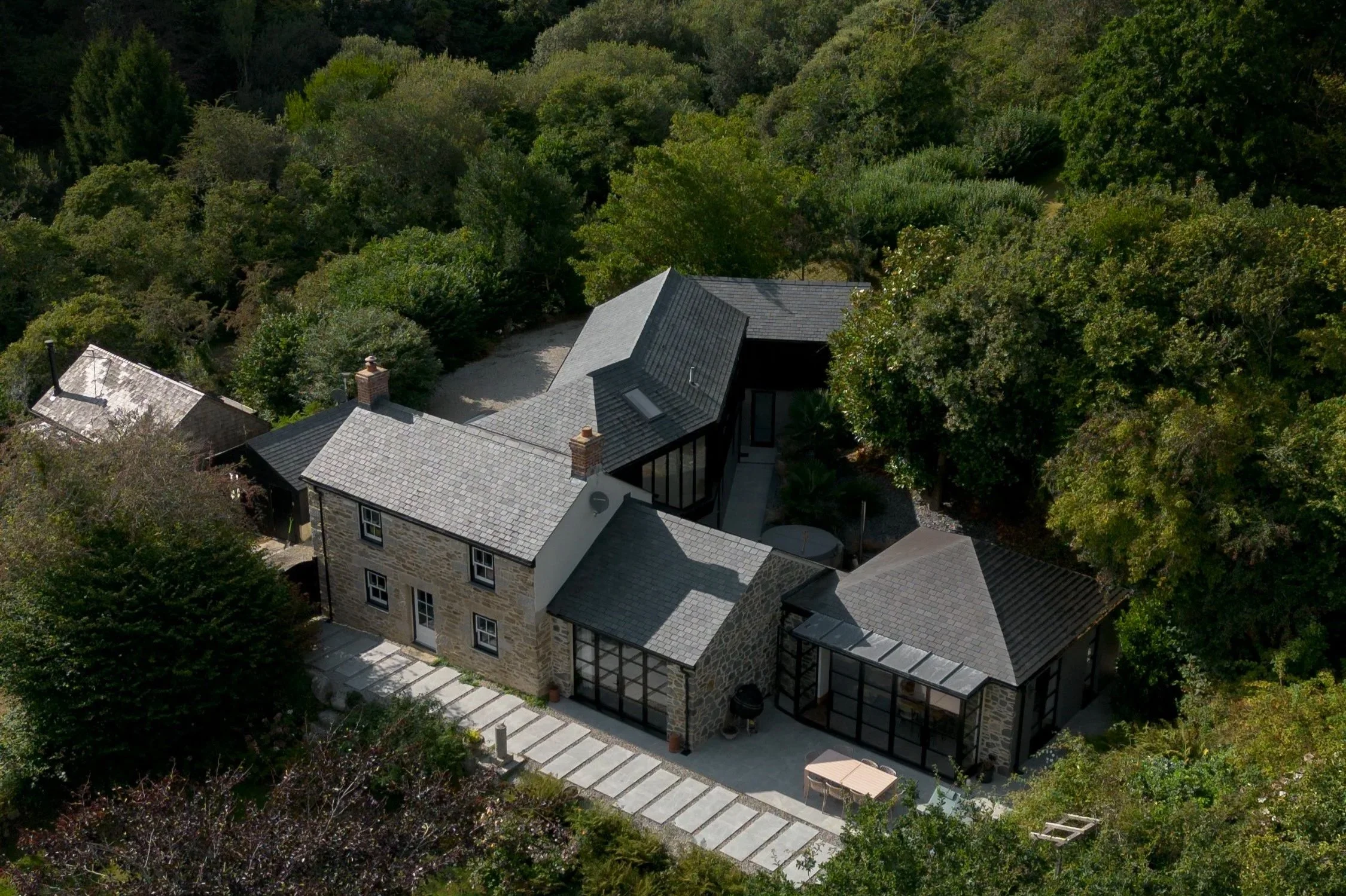 Aerial view of a house surrounded by dense green trees, with a outdoor patio and pathway visible.