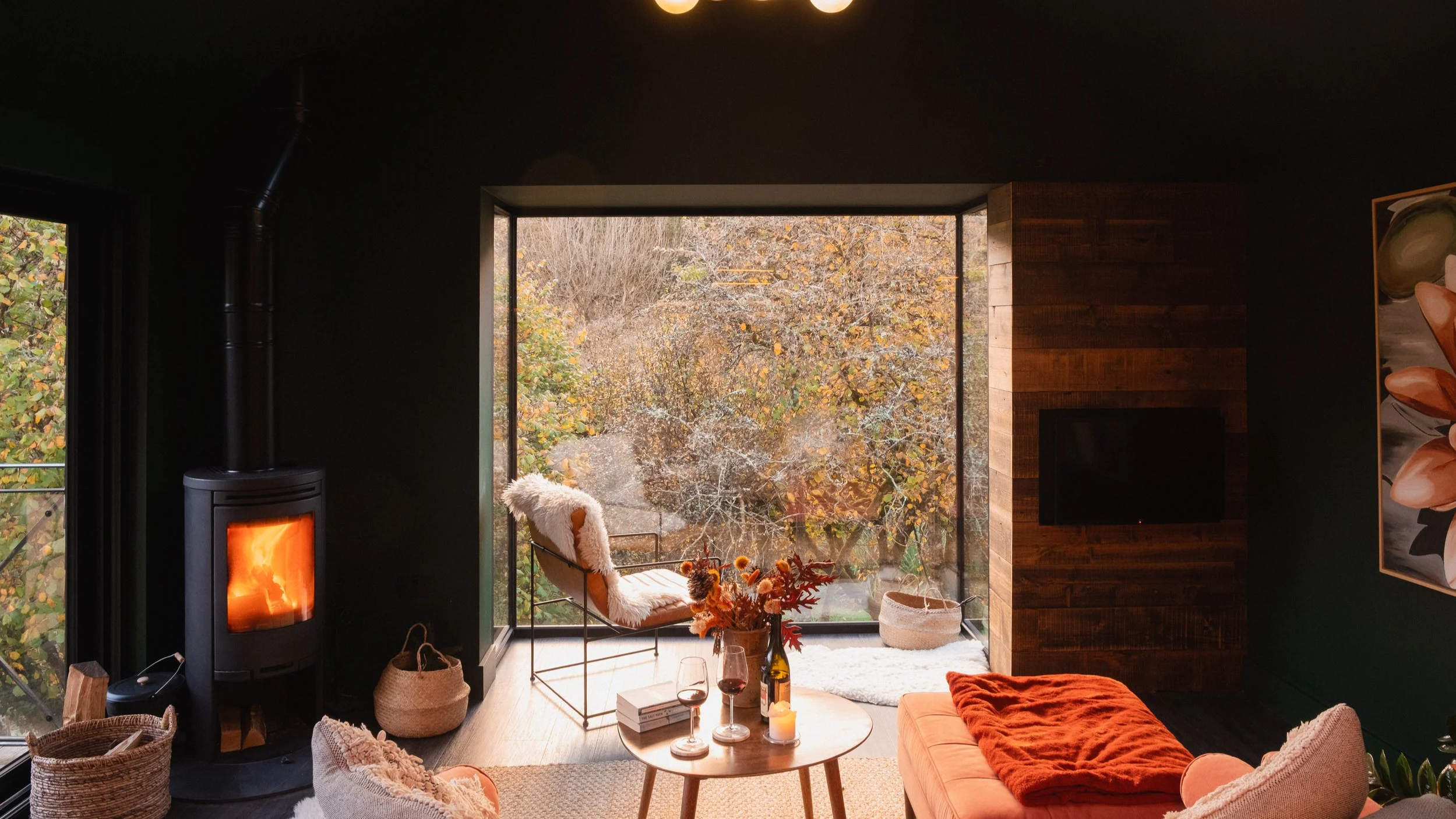 Modern living room with a large window overlooking a fall landscape, a wood-burning stove, cozy chairs with blankets, a small round coffee table with wine glasses and flowers, and a flat-screen TV on a wooden accent wall.