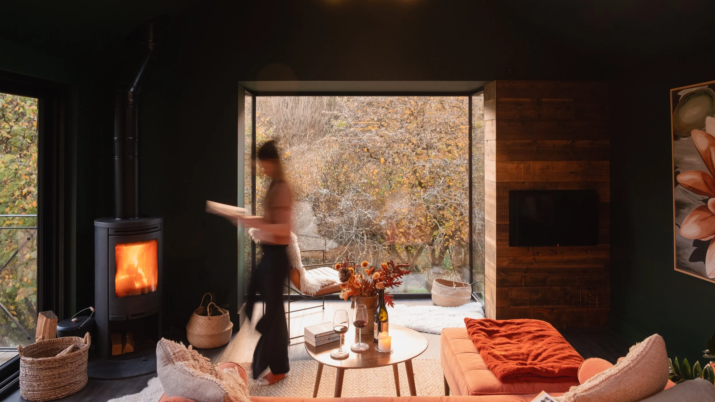 Cozy living room with a woman holding a book near a lit wood stove, large window showing trees, floral artwork, and furniture with cushions and a vase of flowers.