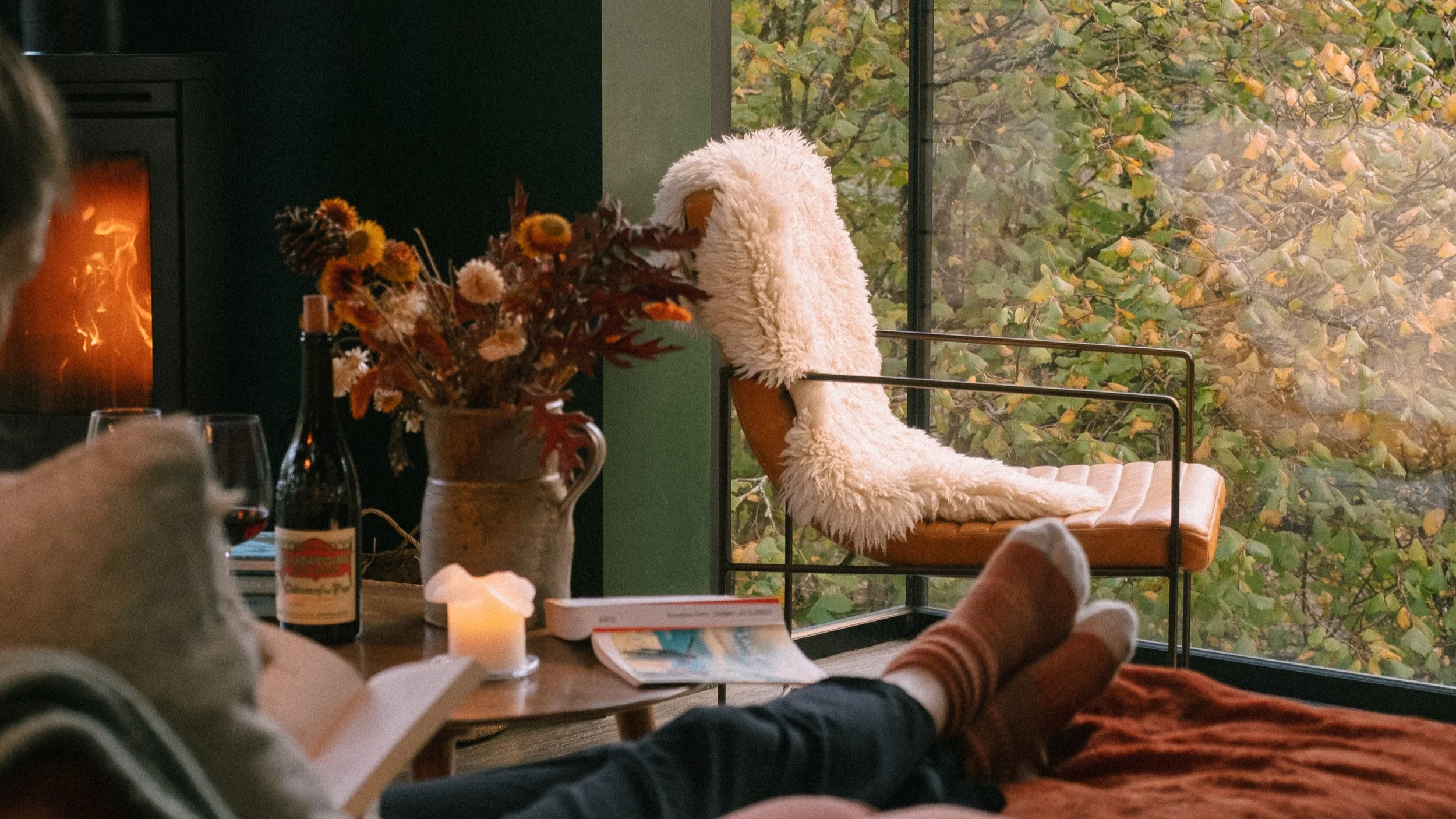 A cozy indoor scene with a person reading a book, a bottle of red wine, and a glass on a wooden table, featuring a lit candle and a large vase of dried flowers. Outside the window, autumn leaves cover the ground.